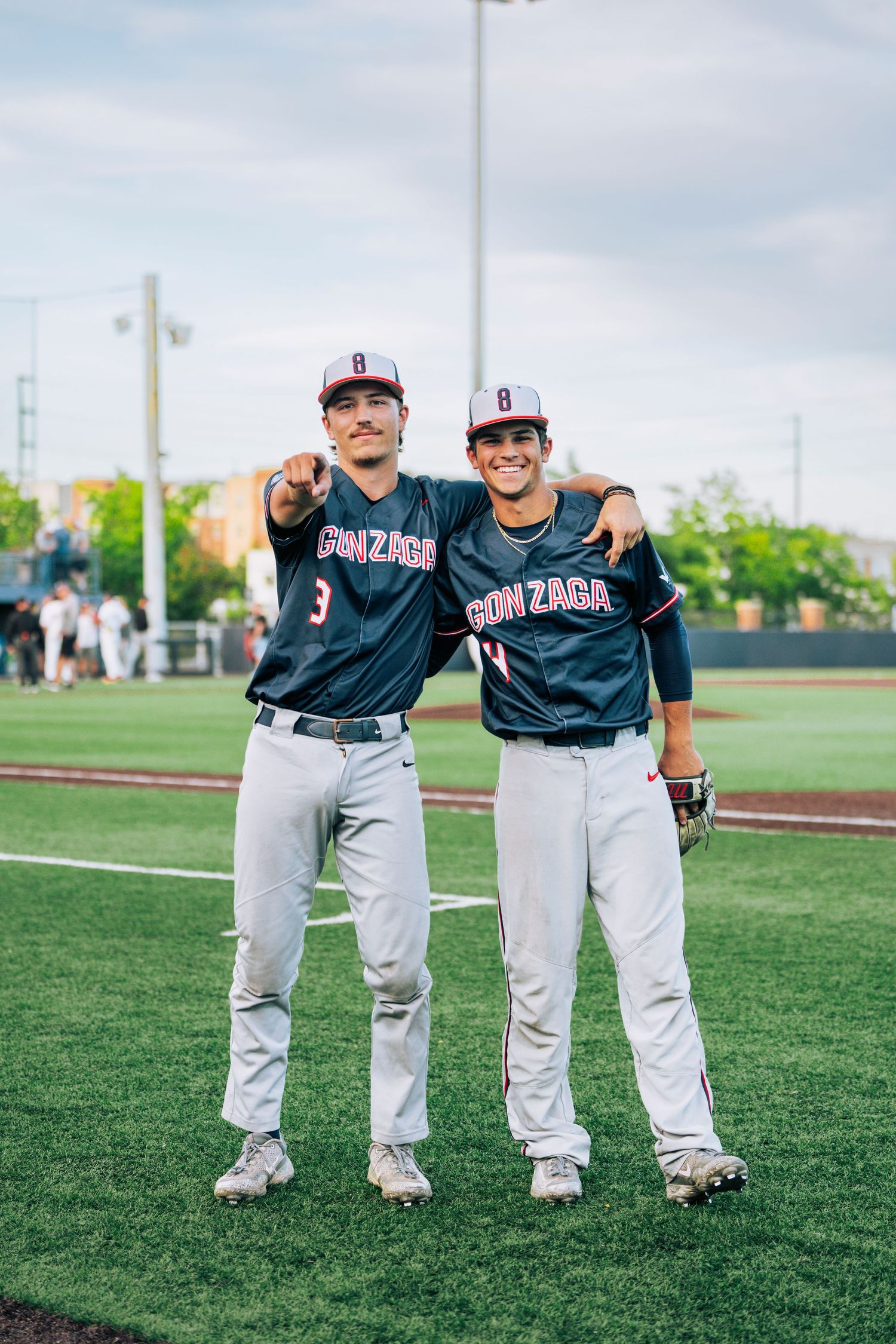 Two baseball players in navy Gonzaga jerseys pose together on a green athletic field under cloudy skies.