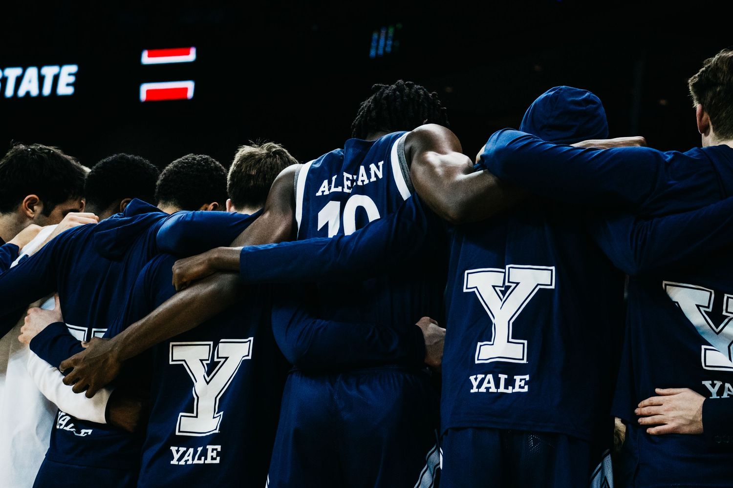 Basketball players huddle together wearing Yale jerseys with arms around each other's shoulders.