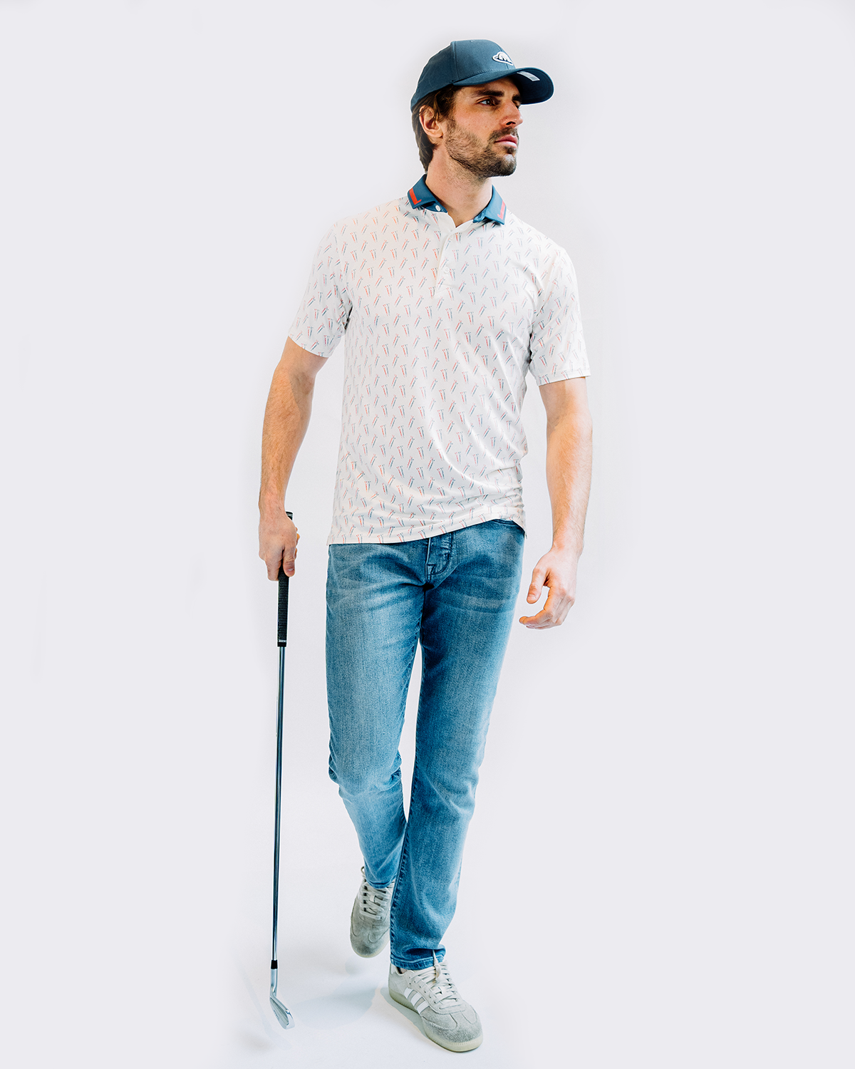 A golfer in casual attire wearing jeans, a white polo shirt and baseball cap stands with a golf club against white background.