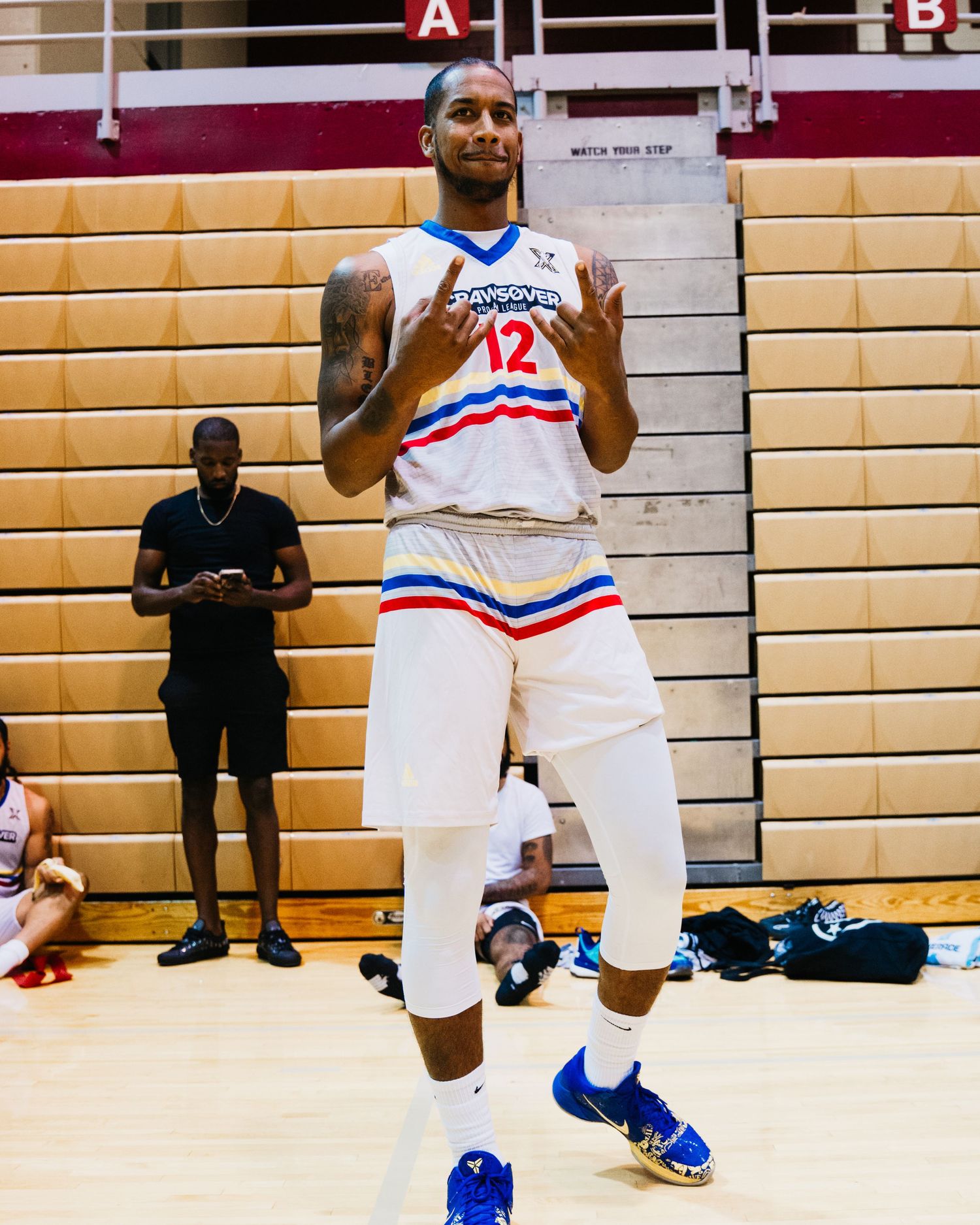 Basketball player in white uniform with red and blue stripes stands on court with arms raised.