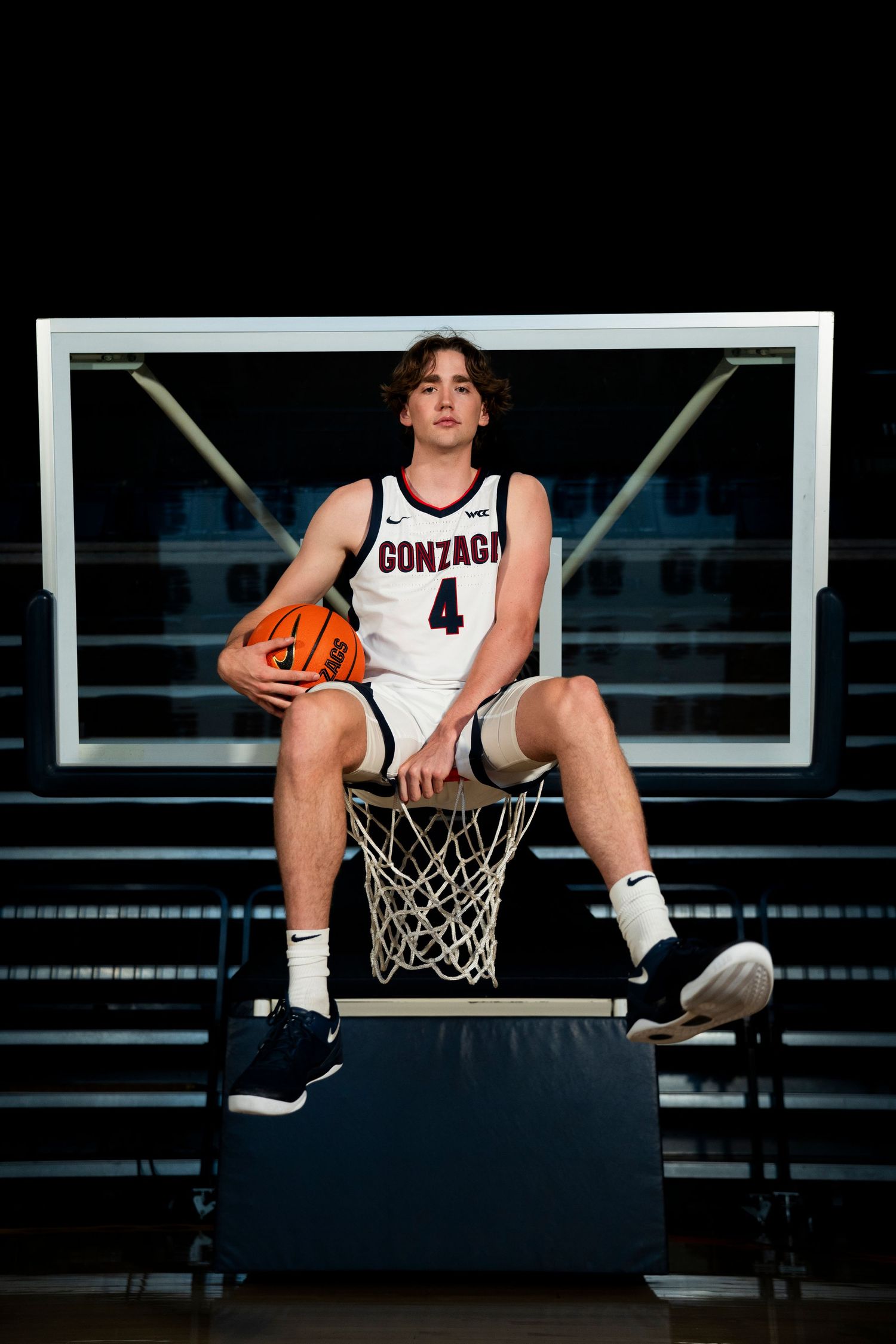 Basketball player in white Gonzaga jersey number 4 sits on basketball hoop rim holding orange ball in dark gym.