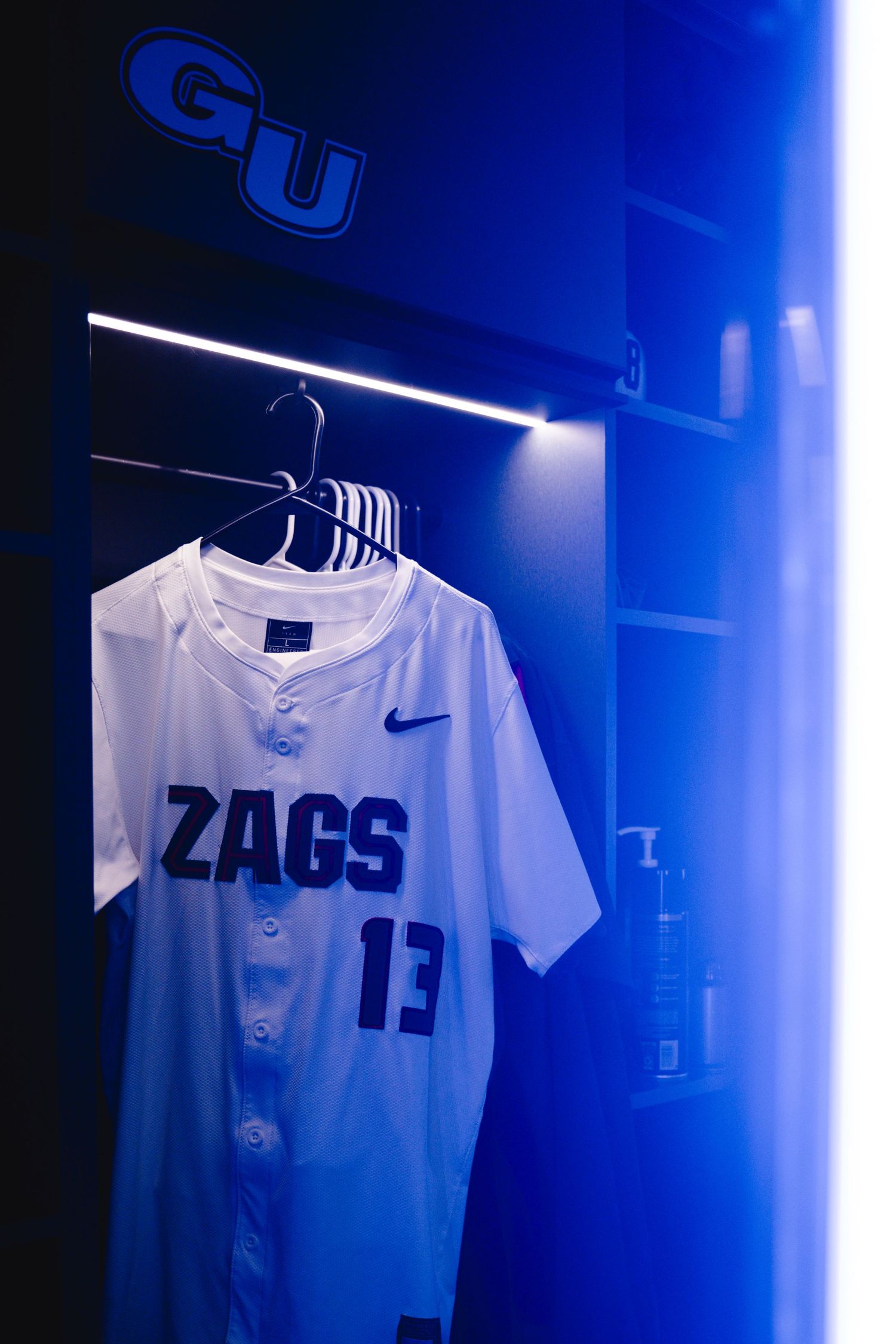 A Gonzaga Bulldogs basketball jersey with number 13 hangs illuminated in blue light against a dark background.