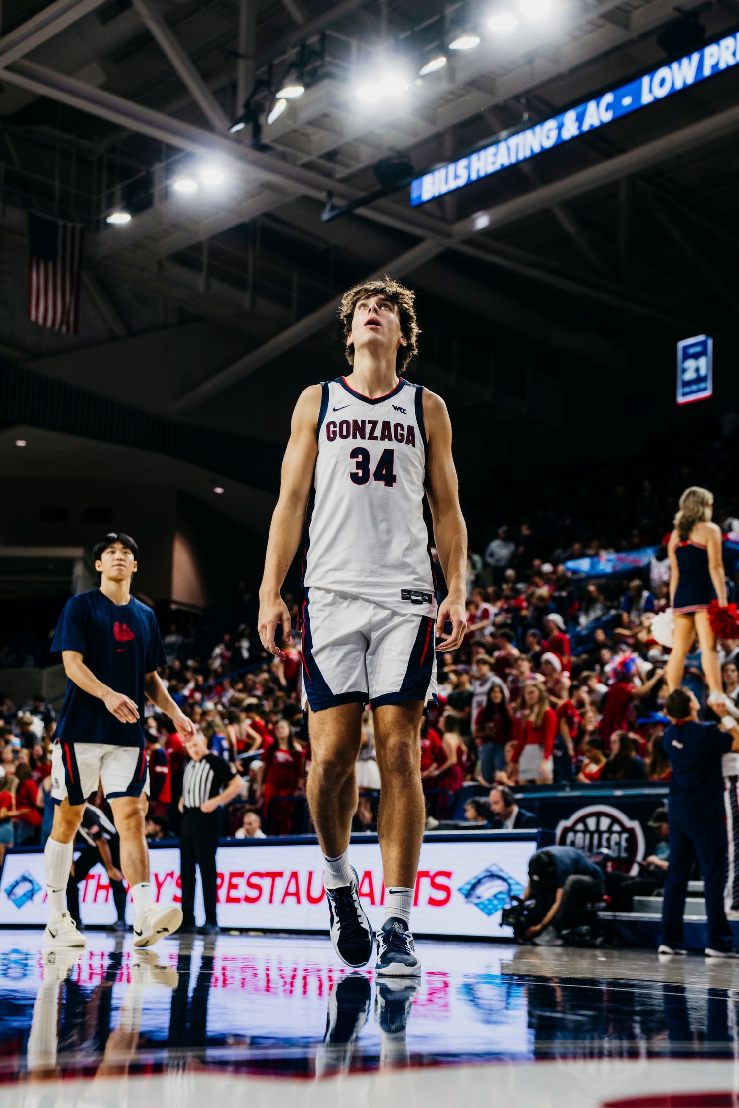 College basketball player in white Gonzaga #34 jersey leaps on court during competitive game under stadium lights.