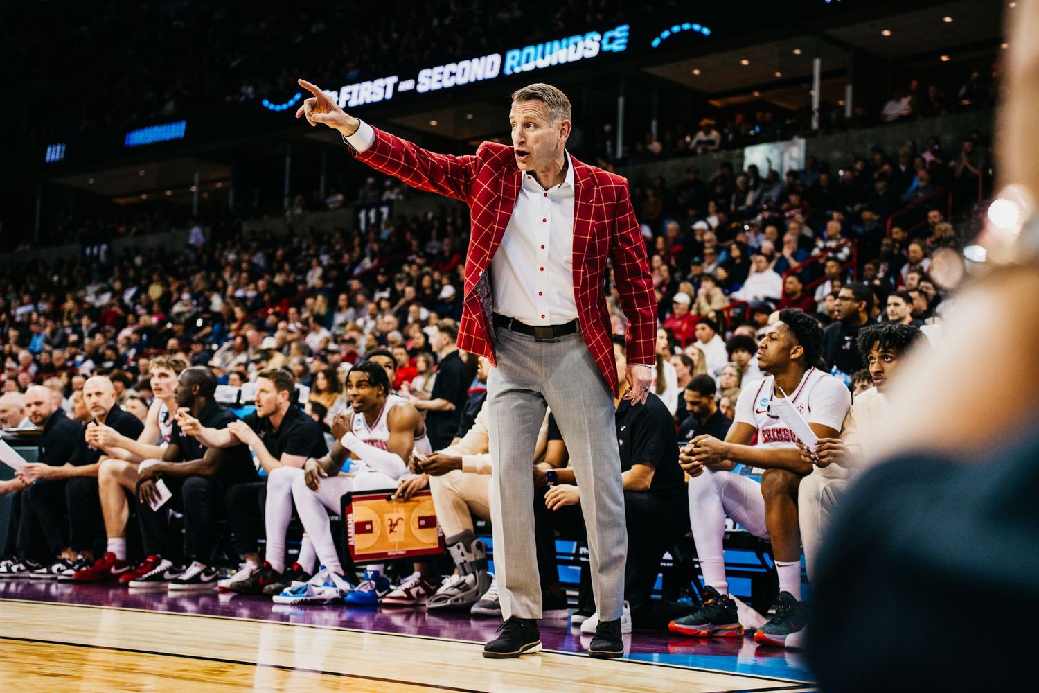 Basketball coach in red jacket gestures animatedly on sideline during game in packed arena.