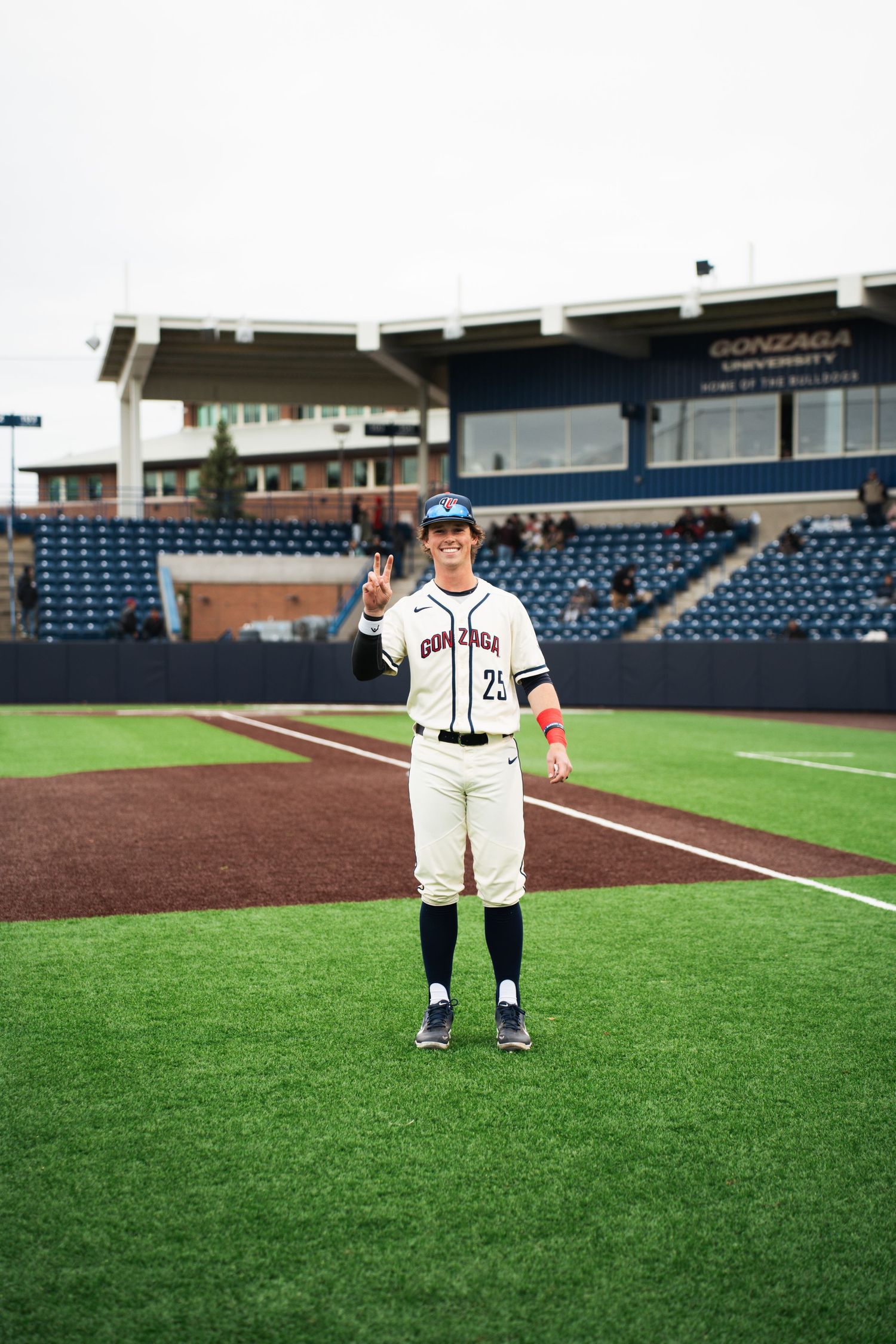 Baseball player in white uniform stands on field giving thumbs up at minor league stadium.
