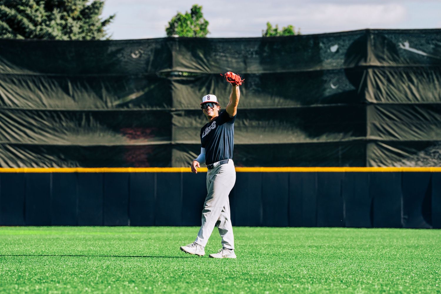 Baseball player in white uniform raises glove high to catch a fly ball on green outfield grass near black wall.