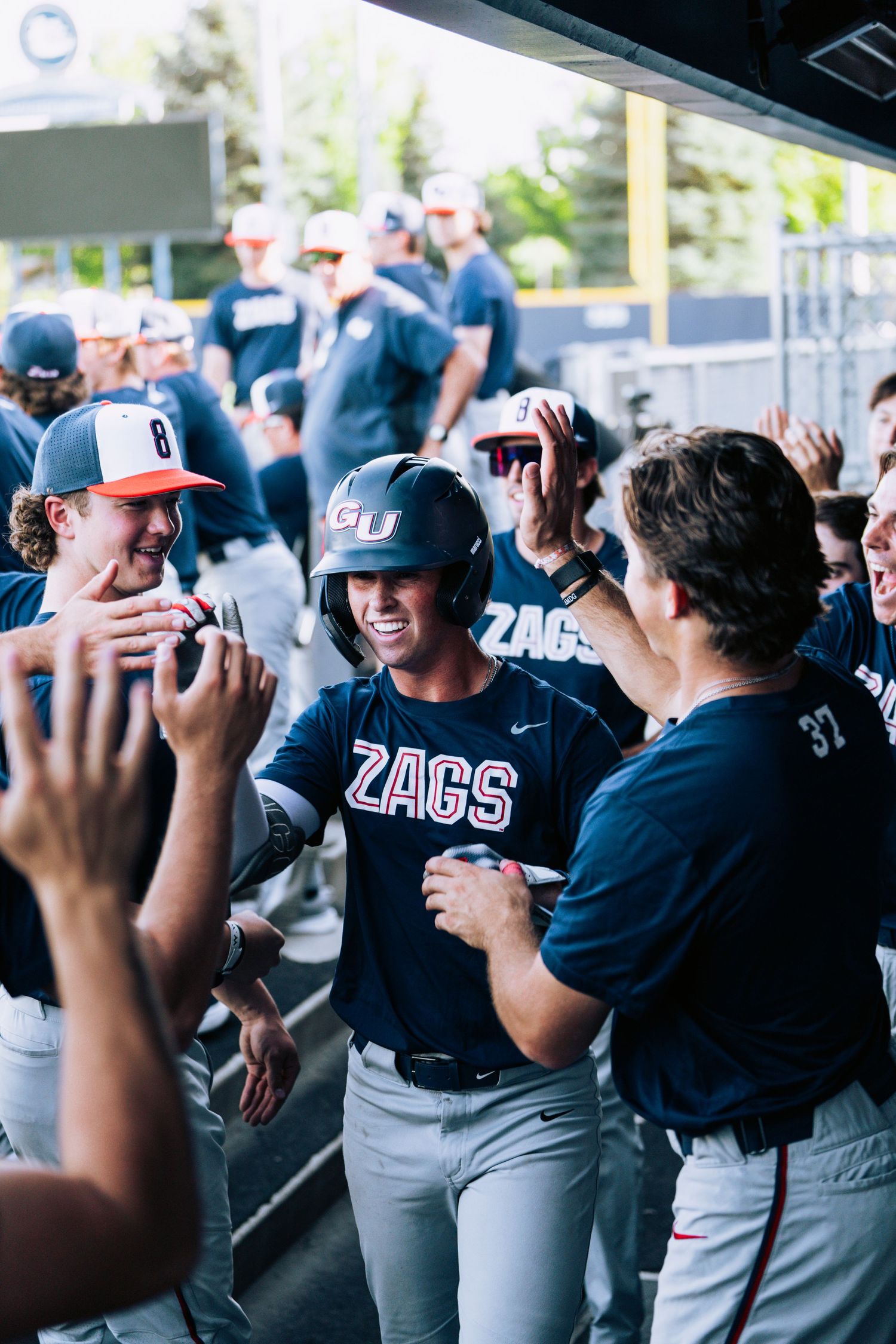 Baseball players in Gonzaga Zags uniforms gather in the dugout during a game, sharing team moments.
