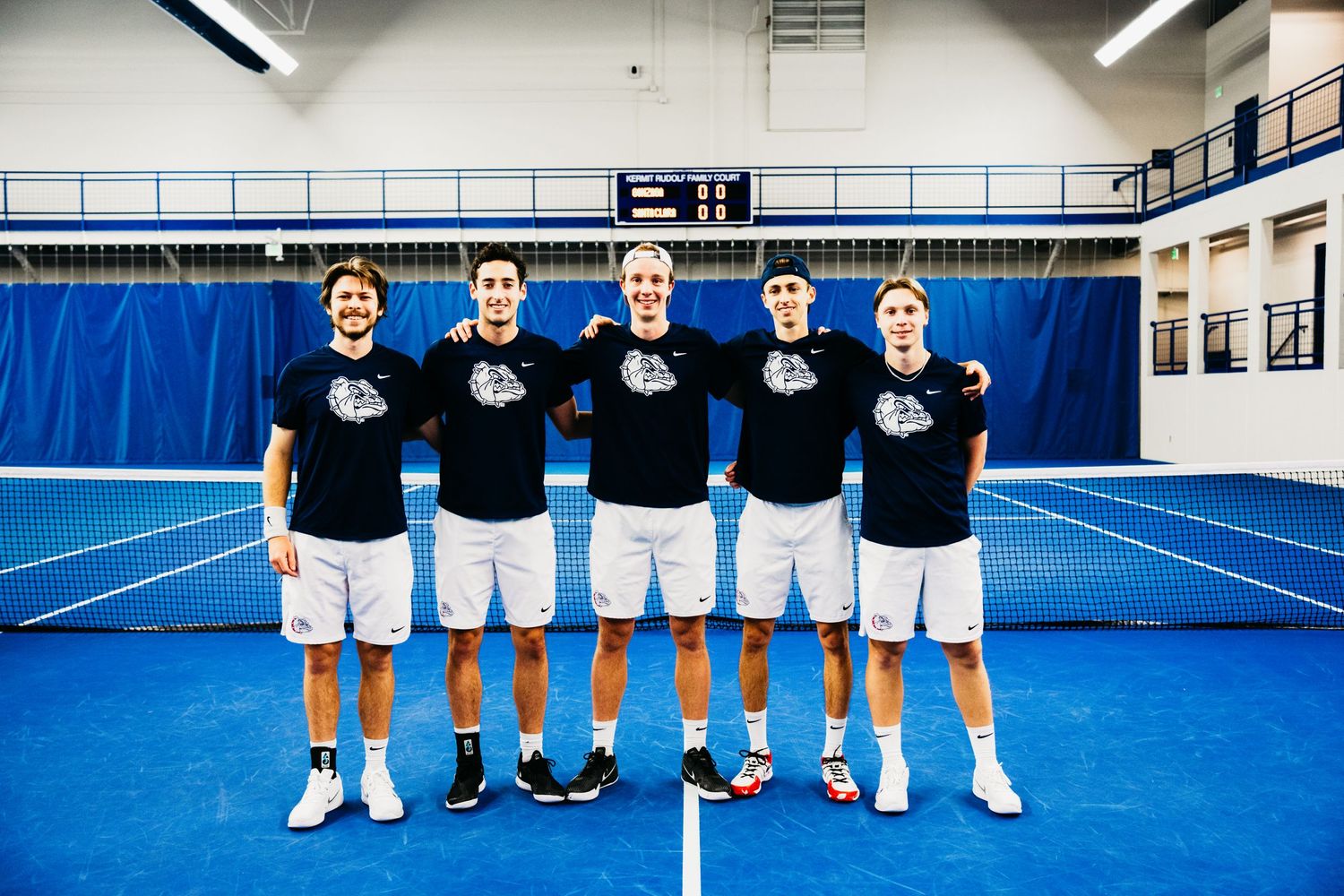 Tennis team members pose on an indoor court wearing matching black shirts, white shorts, and tennis shoes on blue court.