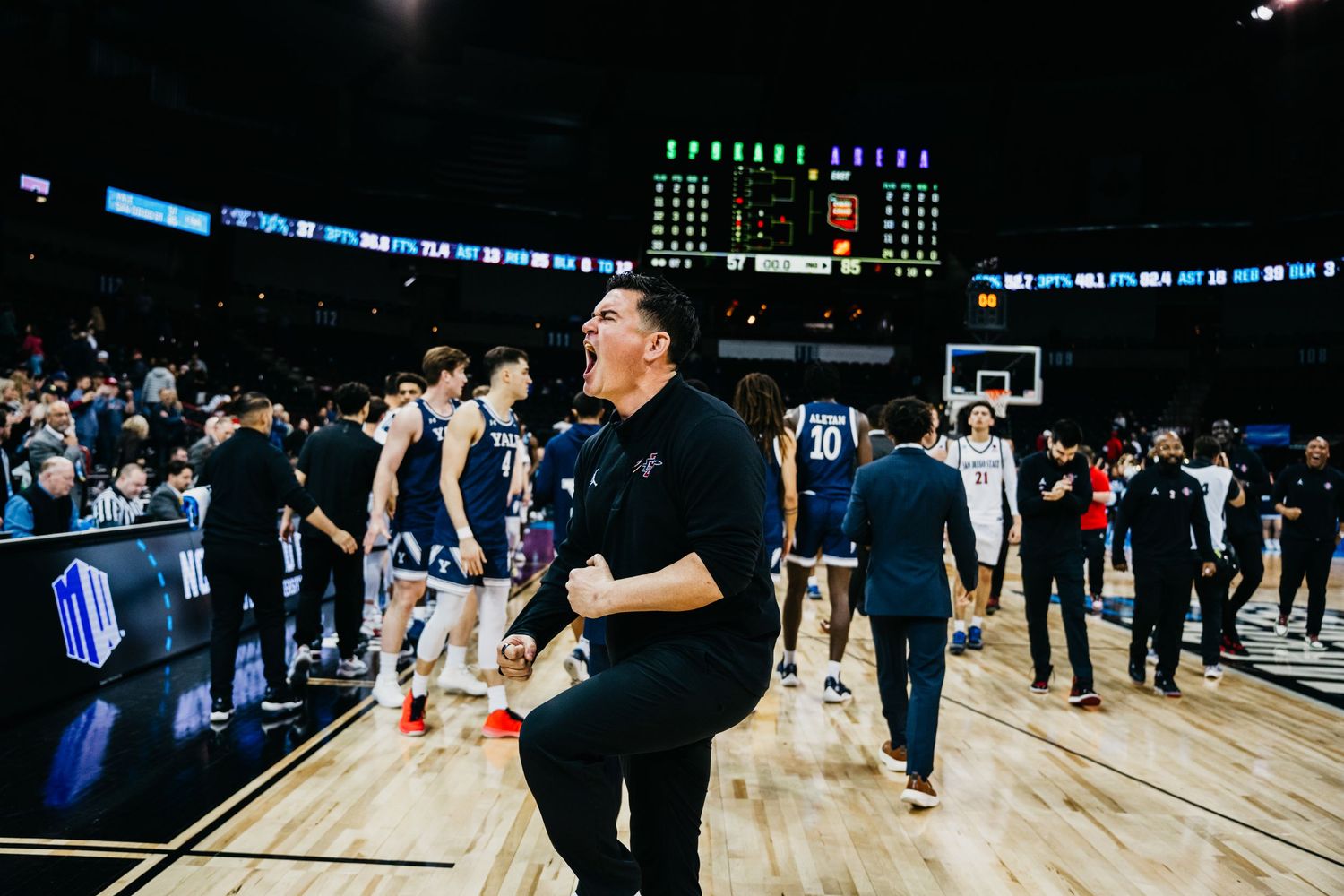 Basketball coach walks on court after game as players and staff gather around the sidelines.