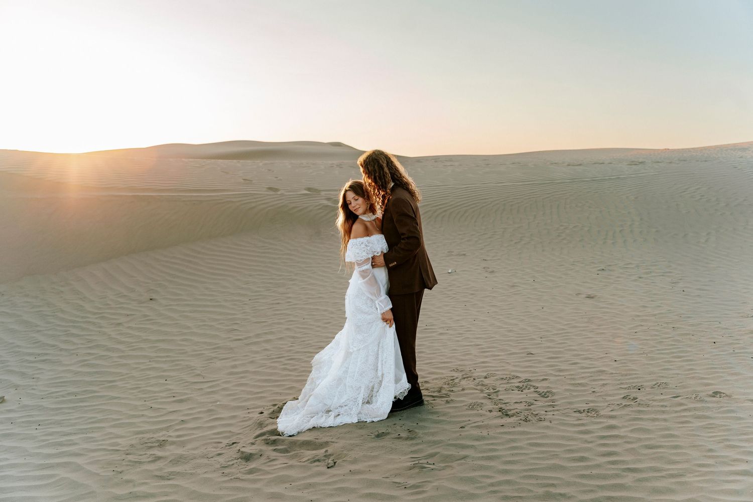 Bride and groom embracing on sandy desert dunes at golden sunset, bride in elegant white mermaid gown.