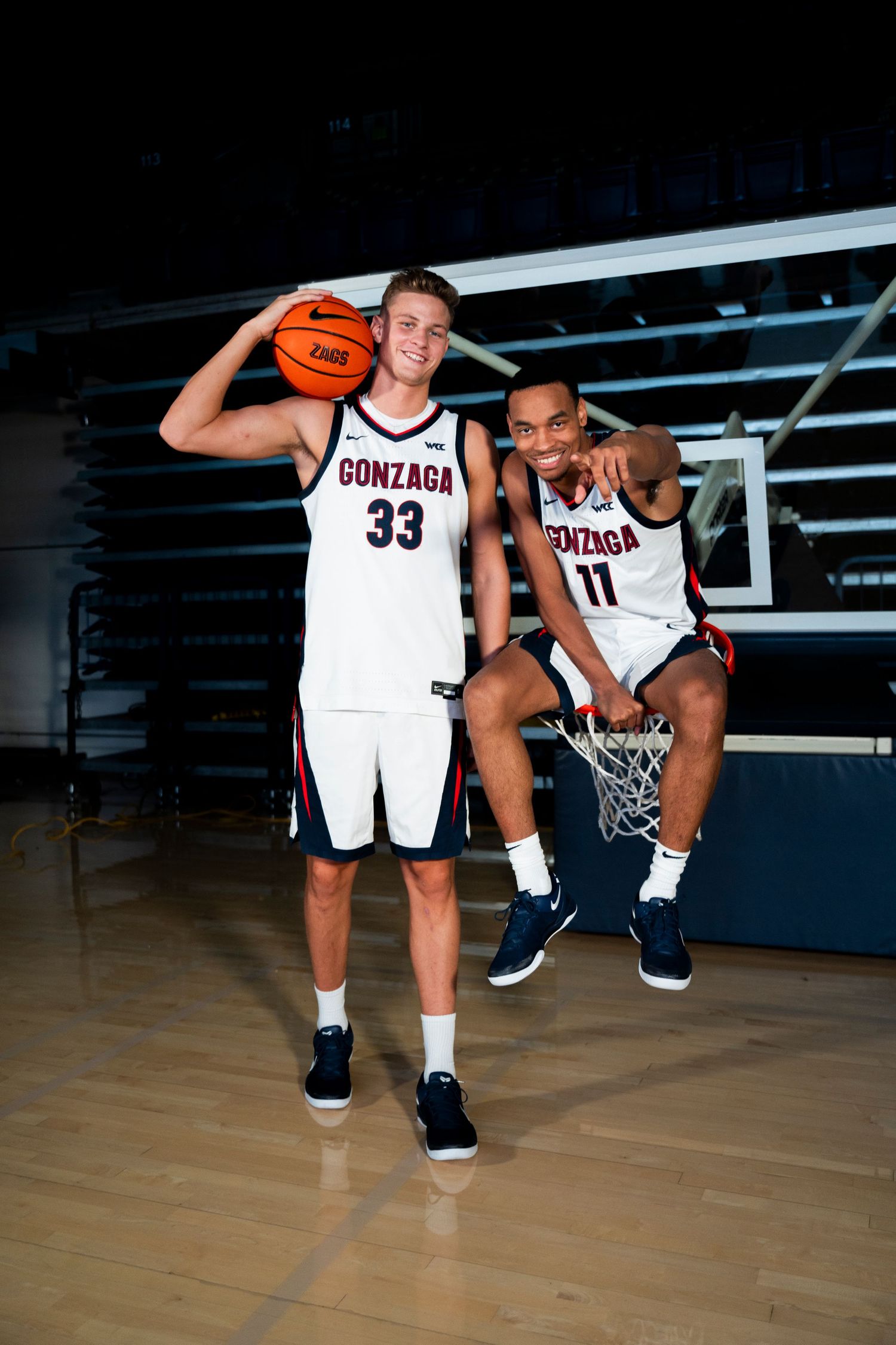 Two Gonzaga basketball players posing on a court, one standing with a ball and one sitting on a hoop in white uniforms.
