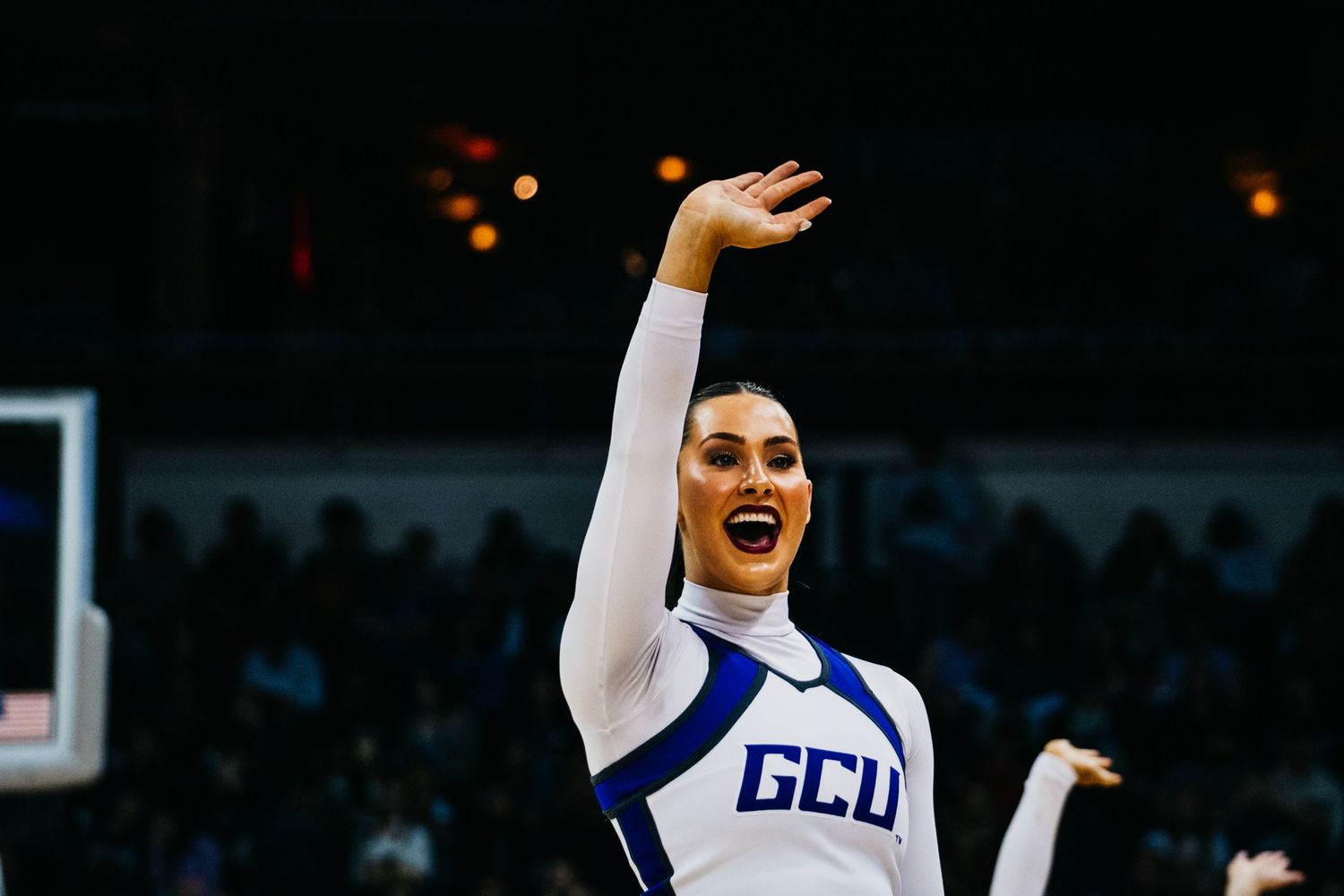 A cheerleader in a white and blue uniform performs energetically with her arm raised on a basketball court.