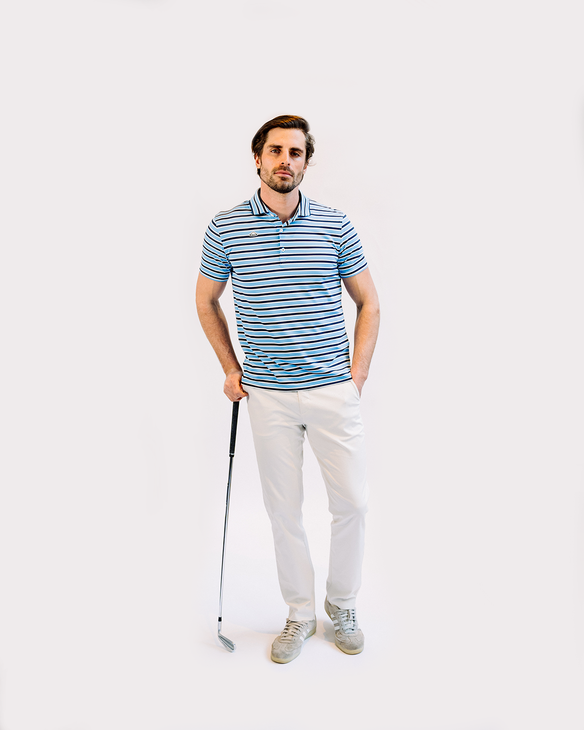Golfer in striped polo shirt and light pants holds club while posing against white background.