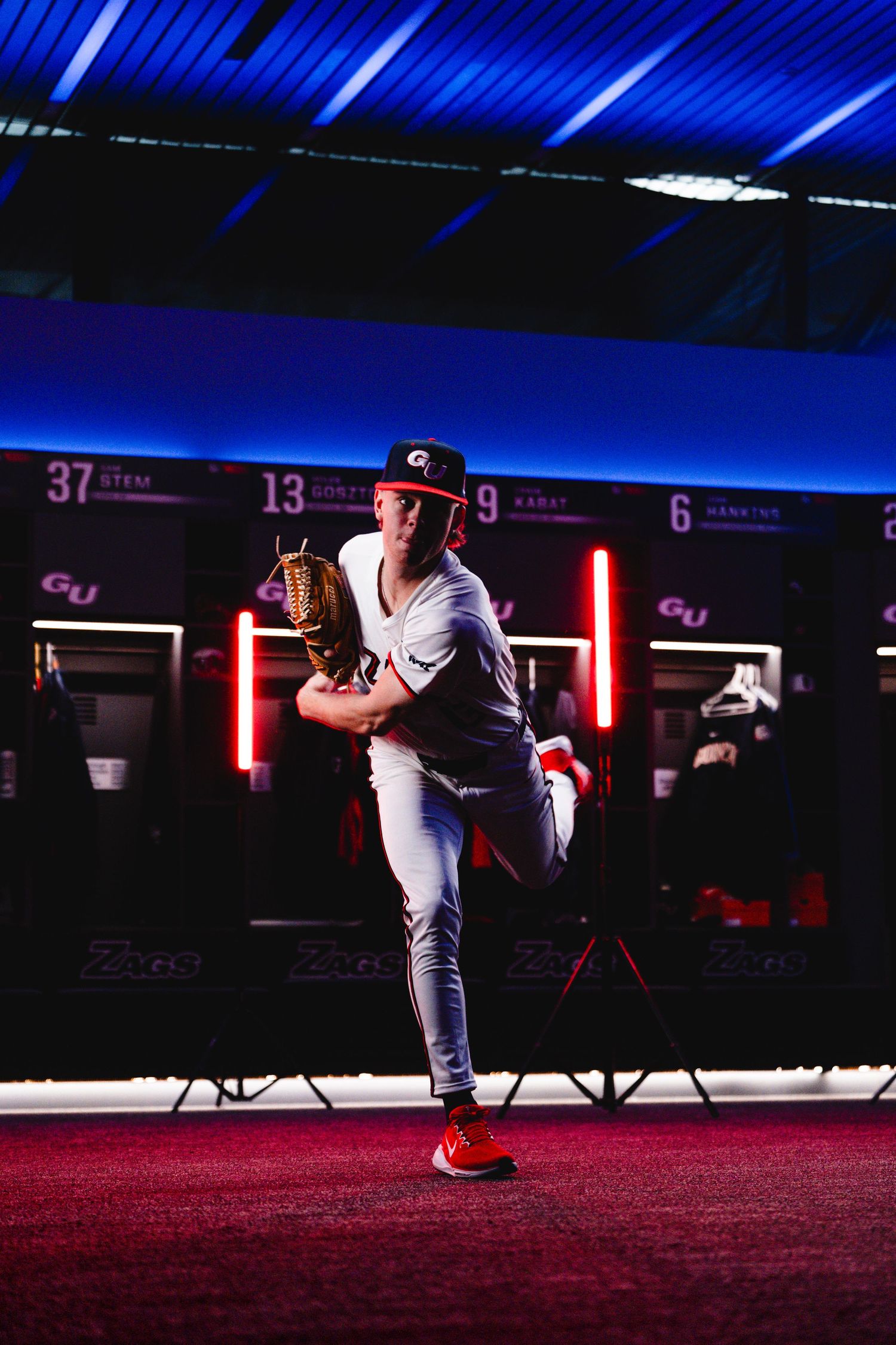 Baseball player in white uniform prepares to pitch in an indoor facility with blue lighting and red neon accents.