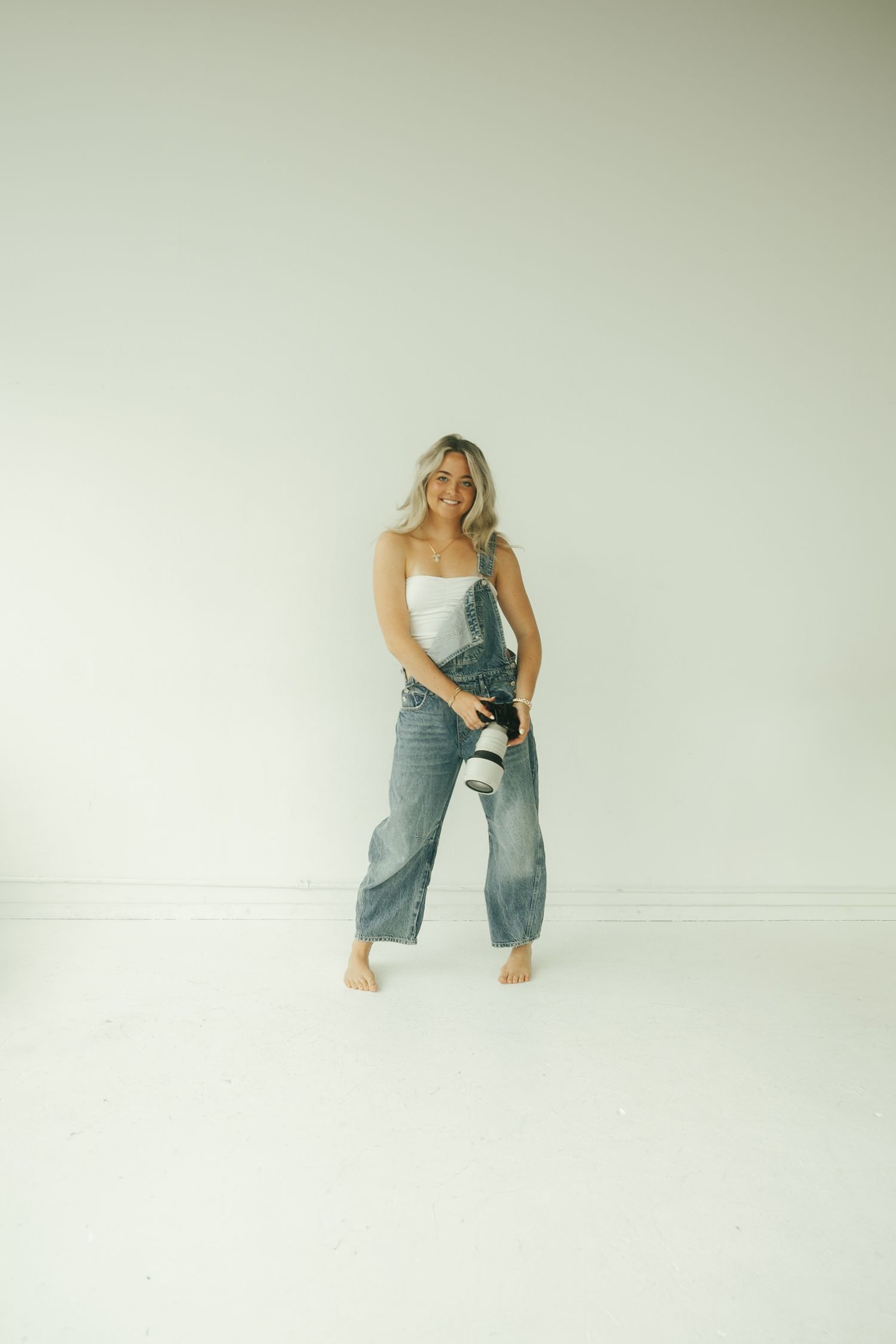 Casual fashion photo of someone wearing denim overalls and white top against minimal white background, standing barefoot.
