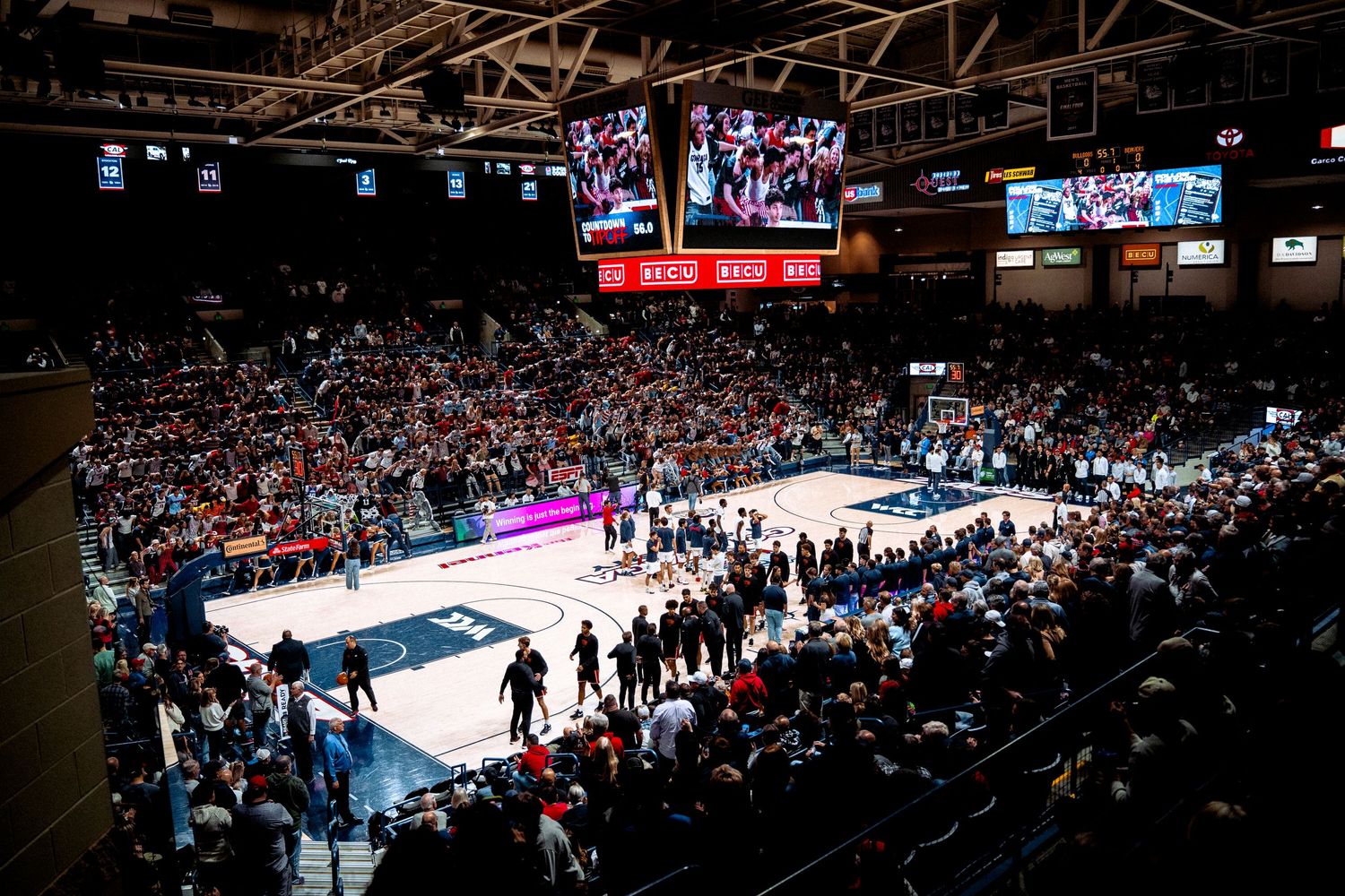 A packed indoor basketball arena with bright lights, video screens and spectators watching a game on the illuminated court.