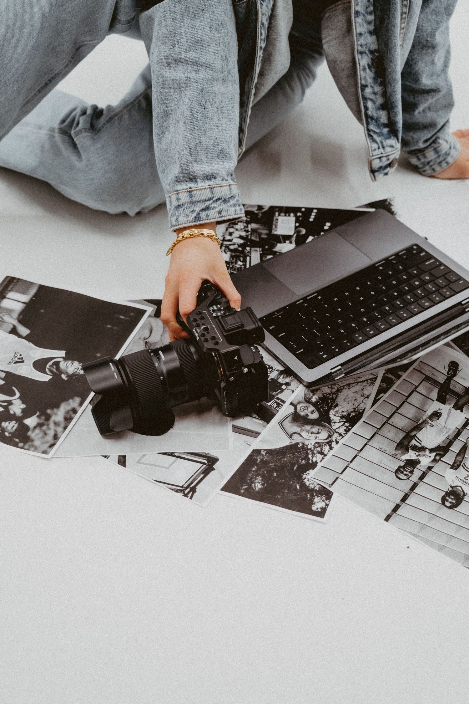 Photographer reviewing black and white prints on floor with DSLR camera and laptop, wearing denim jacket and gold bracelet.