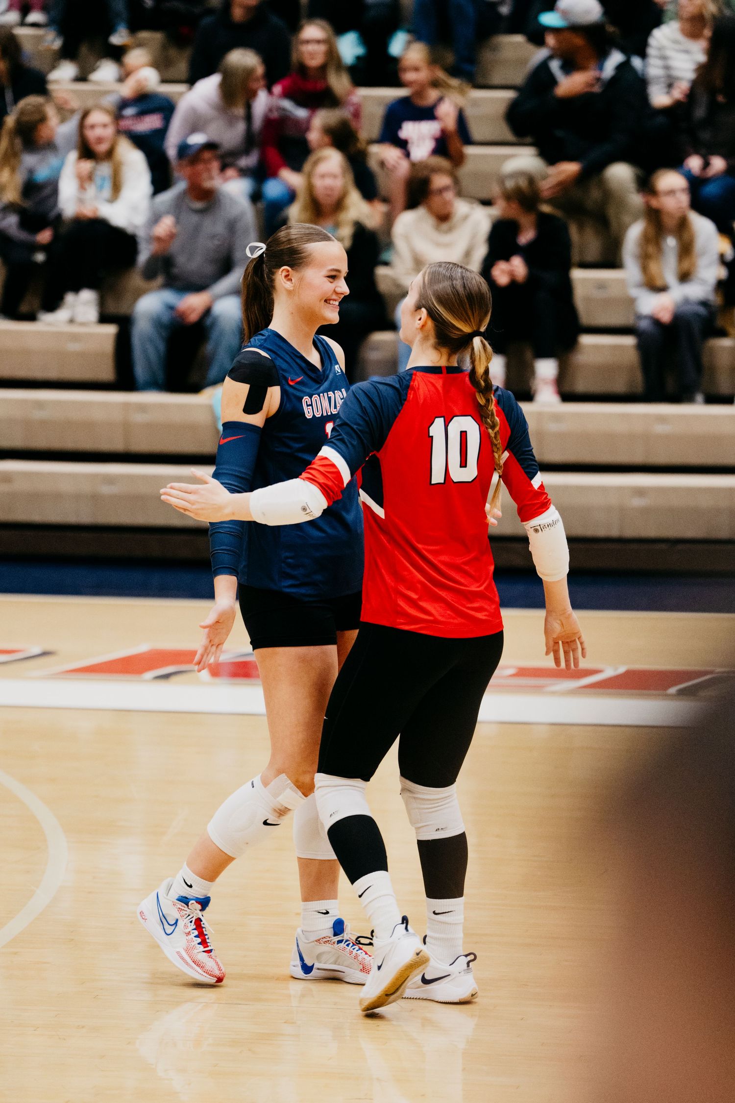 Two volleyball players from opposing teams interact on the court during an indoor match as spectators watch from the bleachers.