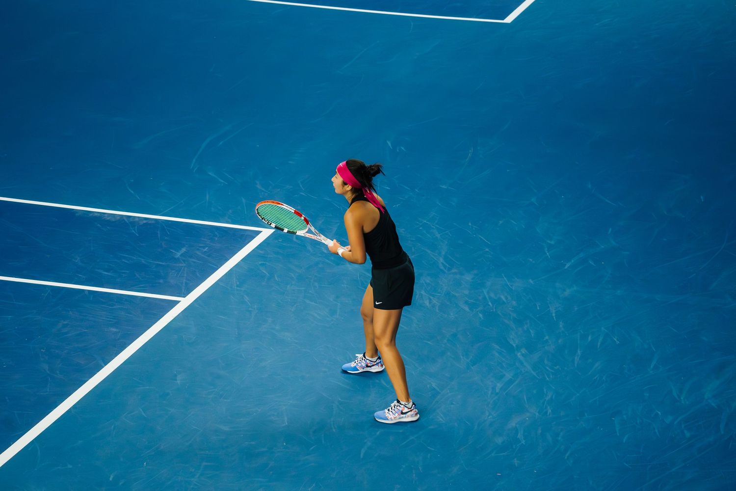 Tennis player in black outfit serves on a bright blue court during a match.