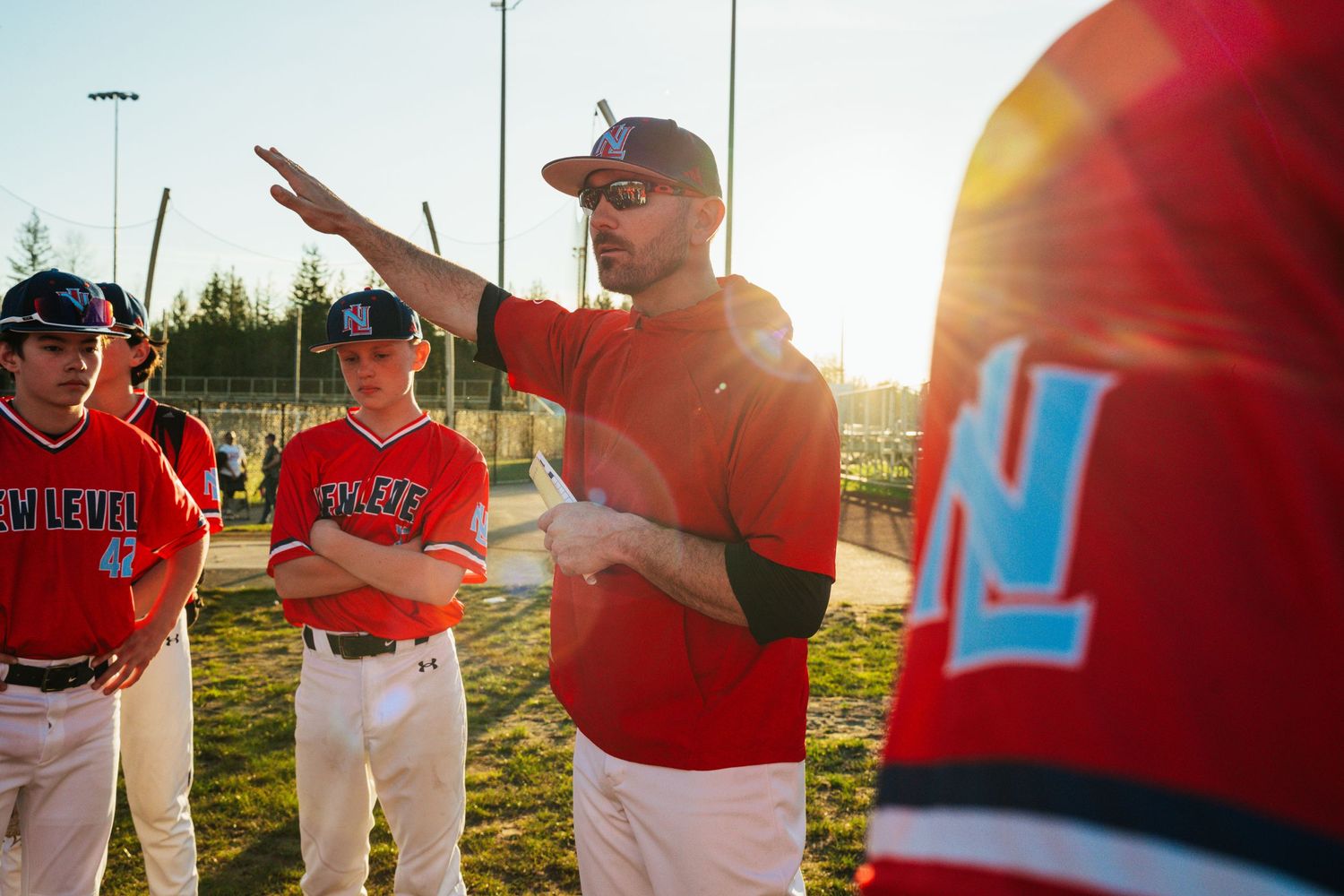 Baseball coach gestures while instructing young players in red uniforms on sunny field.