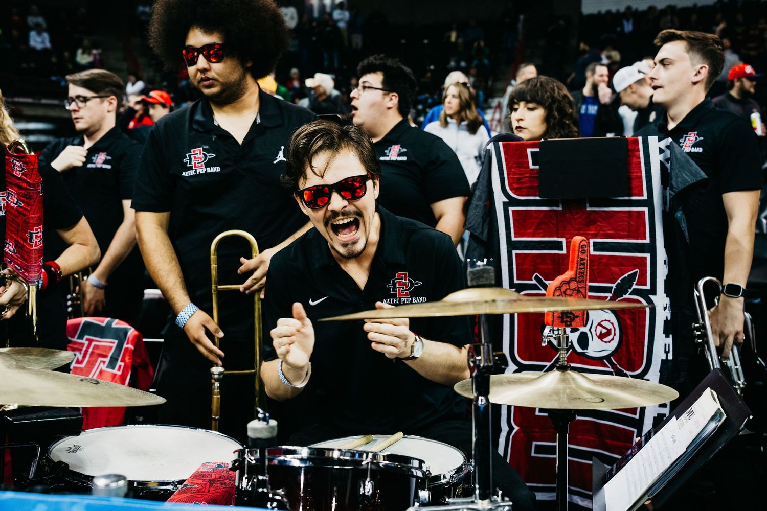 Enthusiastic drummer plays energetically in a crowd at a sporting event or concert, surrounded by other musicians.