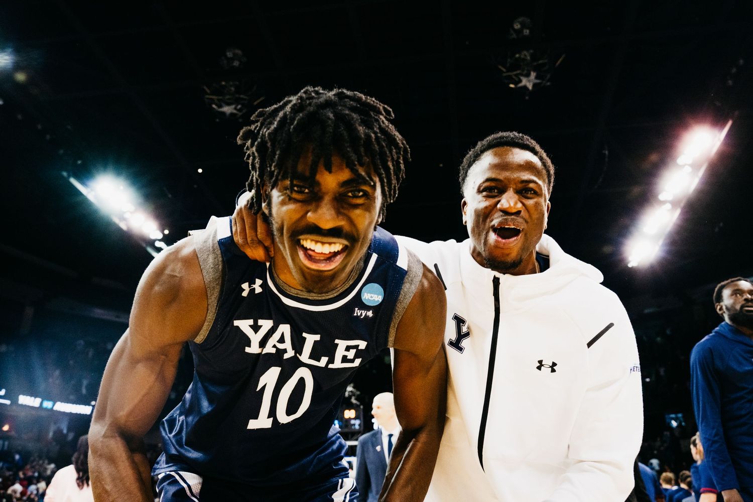 Two smiling basketball players, one wearing a Yale jersey and the other in a white jacket, celebrating after a game.