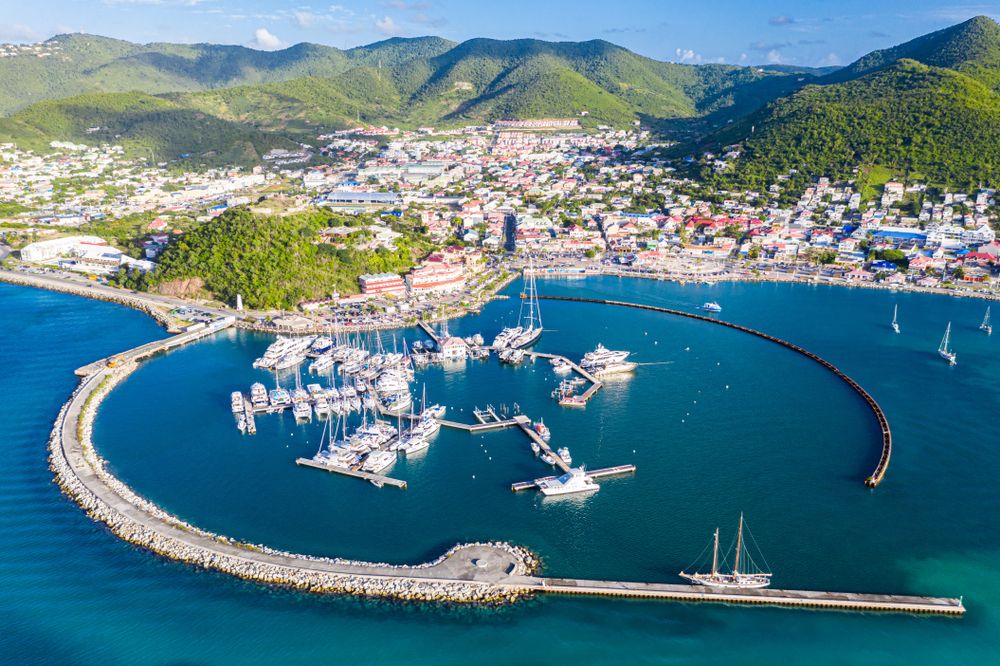 Aerial view of a stunning Caribbean marina with sailboats, curved breakwater, turquoise waters, and colorful hillside town.