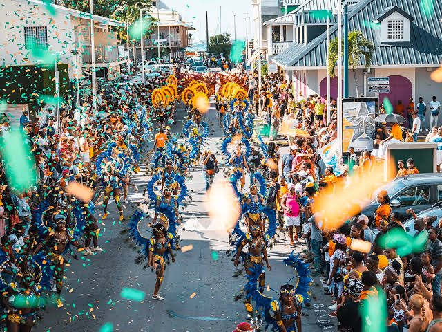 Vibrant Caribbean carnival parade with performers in blue and orange feathered costumes dancing through confetti-filled streets.
