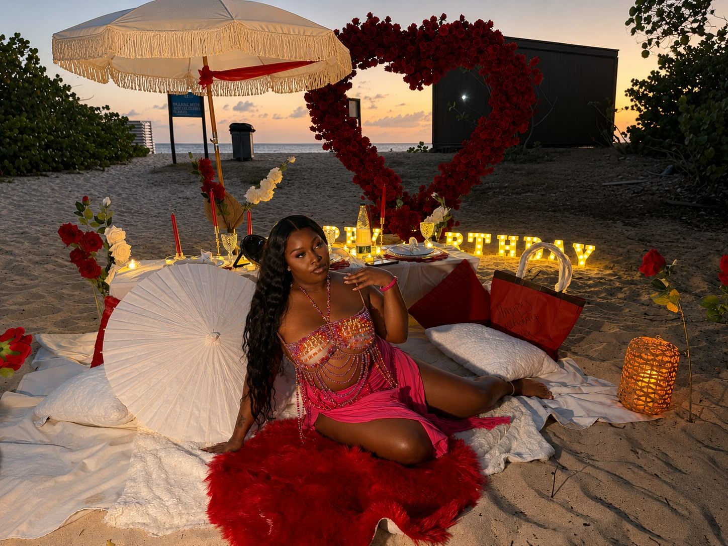 Woman in pink outfit poses on beach birthday setup with red roses, heart decor, and Happy Birthday sign at sunset.