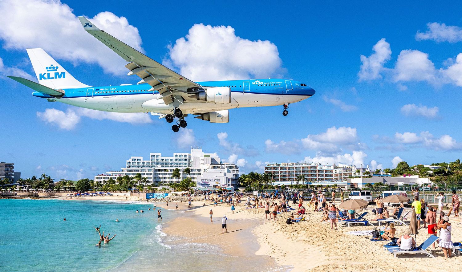 KLM airplane flying low over crowded Maho Beach in St. Maarten with tourists watching the landing.