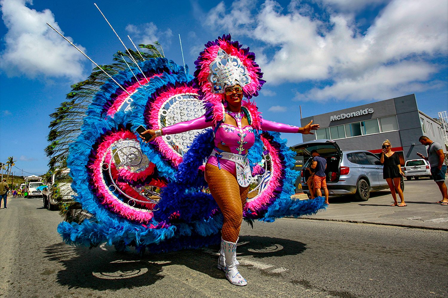 Vibrant carnival dancer in colorful blue and pink feathered costume performing in a street parade under blue skies.