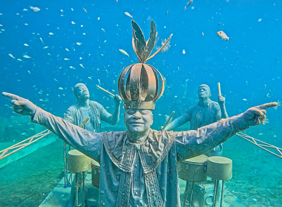 Colorful underwater statues of musicians with a crowned figure and outstretched arms surrounded by tropical fish.