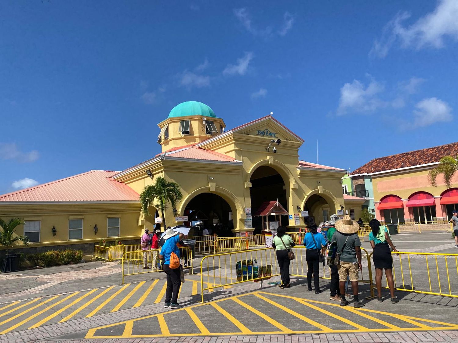Colorful Caribbean shopping center with yellow facade, teal dome, and tourists exploring outside under a bright blue sky.