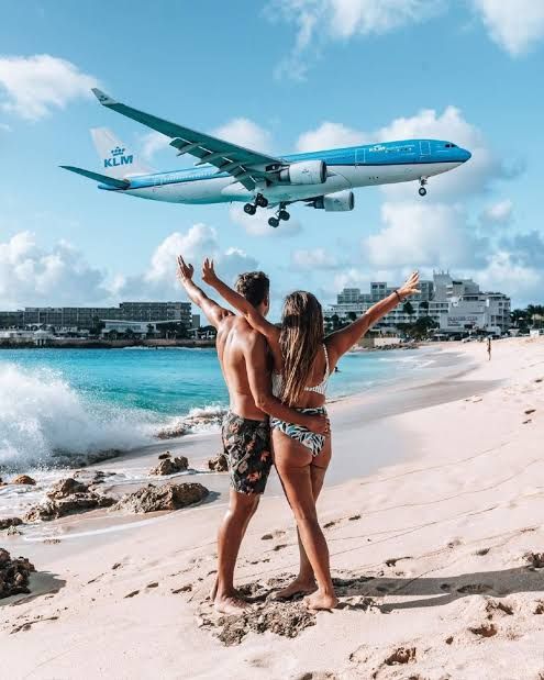 Couple on beach waving at low-flying KLM airplane over Maho Beach, Sint Maarten, turquoise water behind.