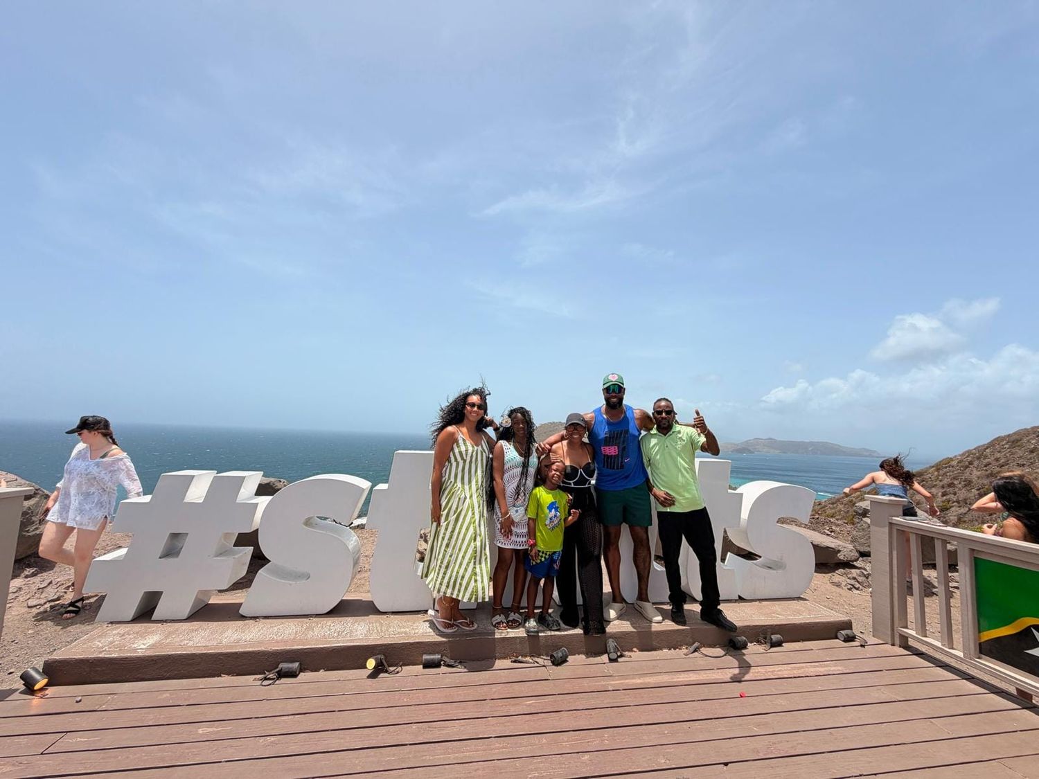 Group of friends posing by a large hashtag sign on a scenic coastal overlook under a bright blue sky.