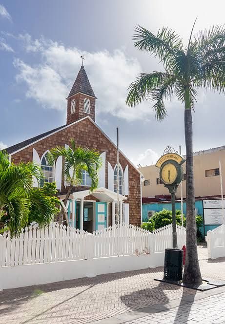 A historic stone church with a white picket fence, palm tree, and vintage clock in a sunny Caribbean town square.