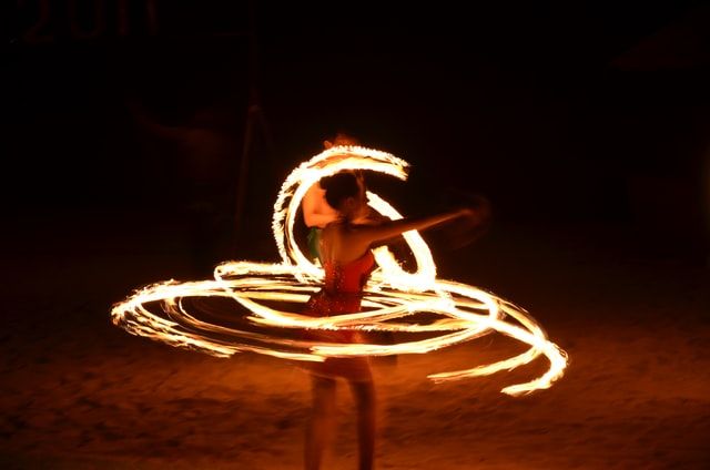 Fire dancer performing at night, creating glowing light trails with spinning fire poi against dark background.