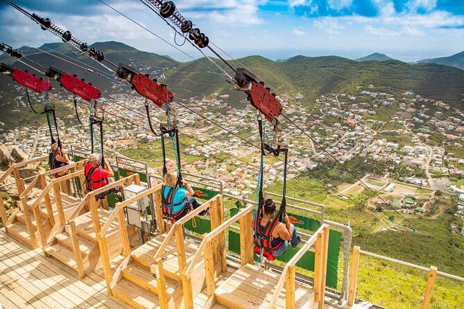 Tourists ride a scenic zip line attraction overlooking a lush mountain valley and coastal Caribbean town below.