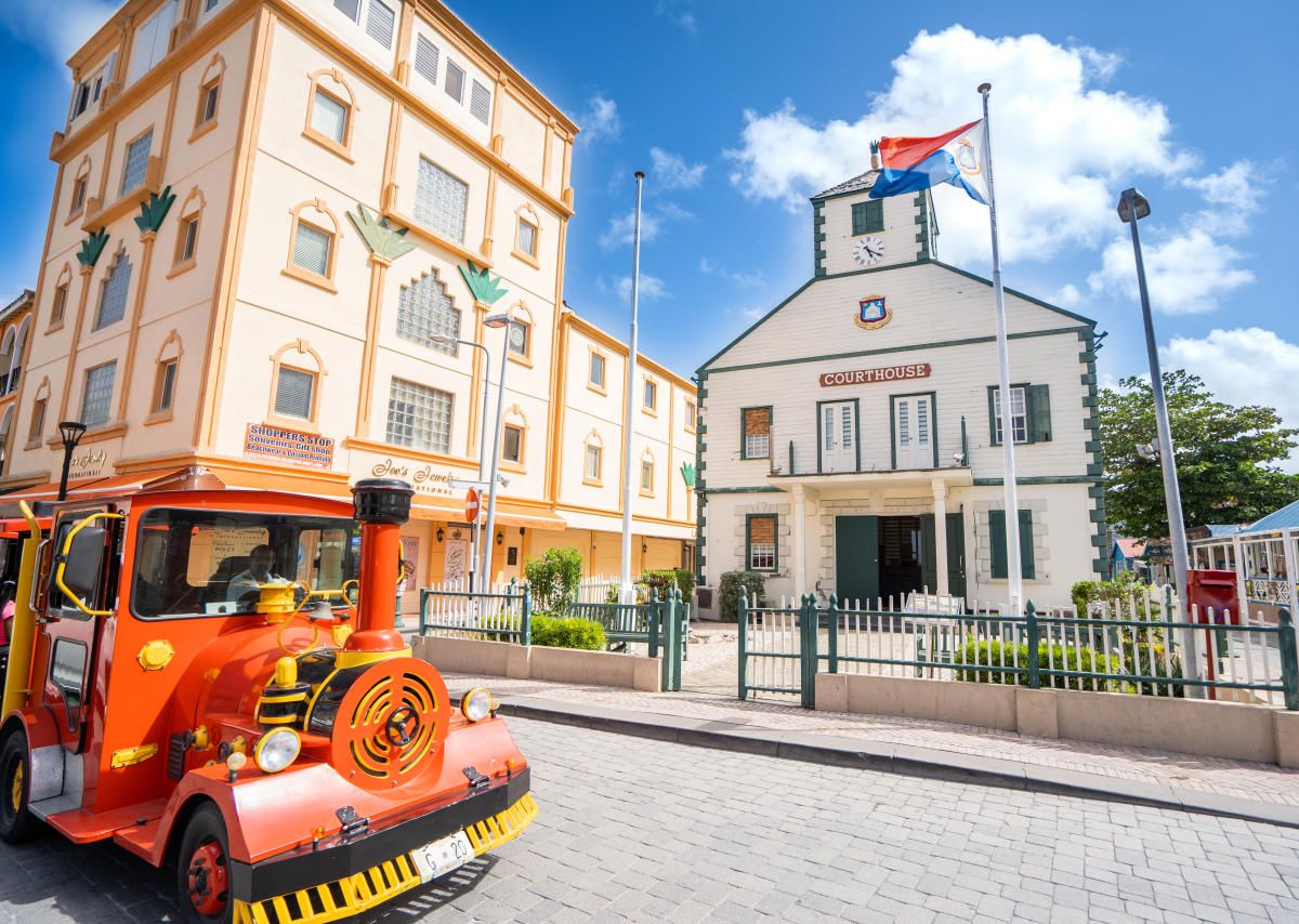 Orange tourist train parked near a historic government building with flags in a sunny Caribbean town square.
