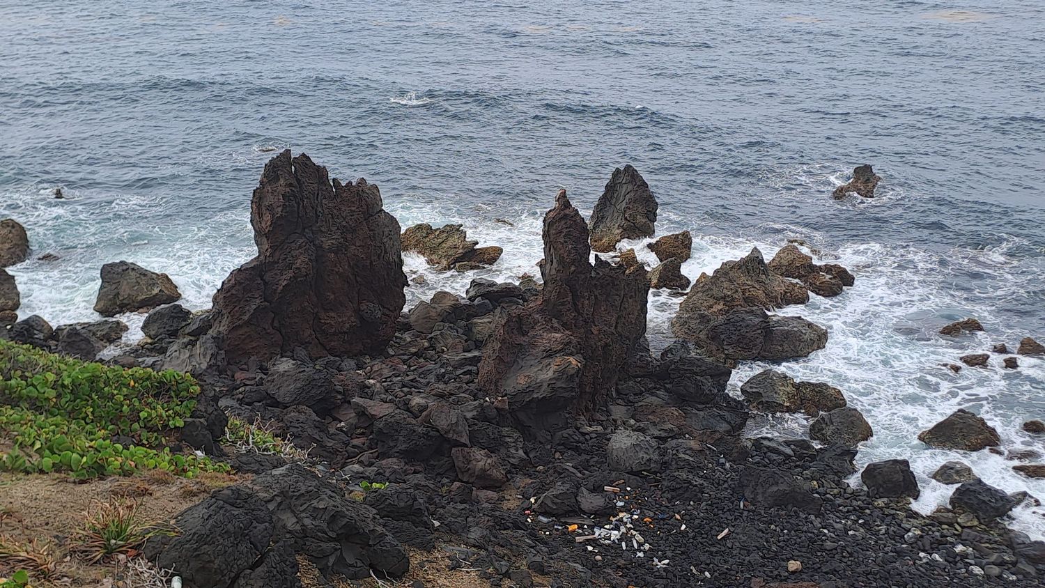 Rocky volcanic coastline with crashing ocean waves and jagged lava rock formations along a rugged shoreline.