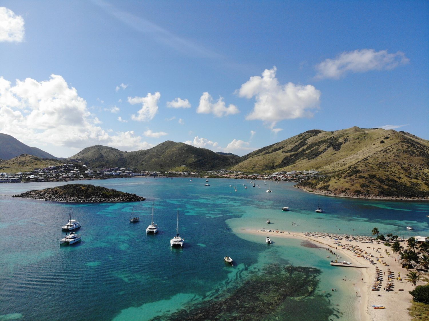 Aerial view of a turquoise Caribbean bay with sailboats, white sandy beach, and lush green hills under a blue sky.