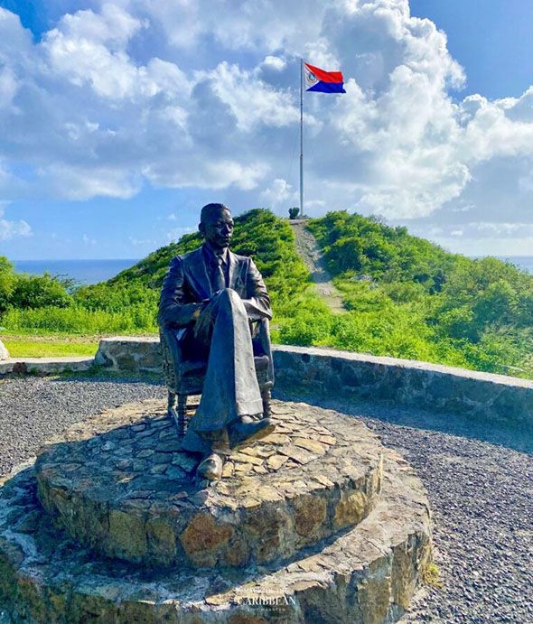 Bronze statue of a seated figure on a stone pedestal with a flag and green hills overlooking the sea.