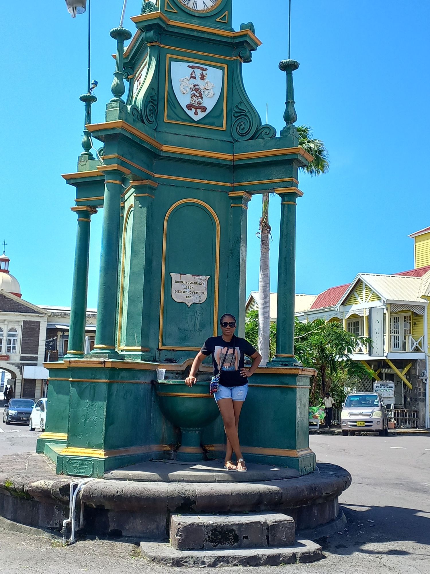 Woman posing by a green Victorian clock tower landmark in a sunny Caribbean town street.