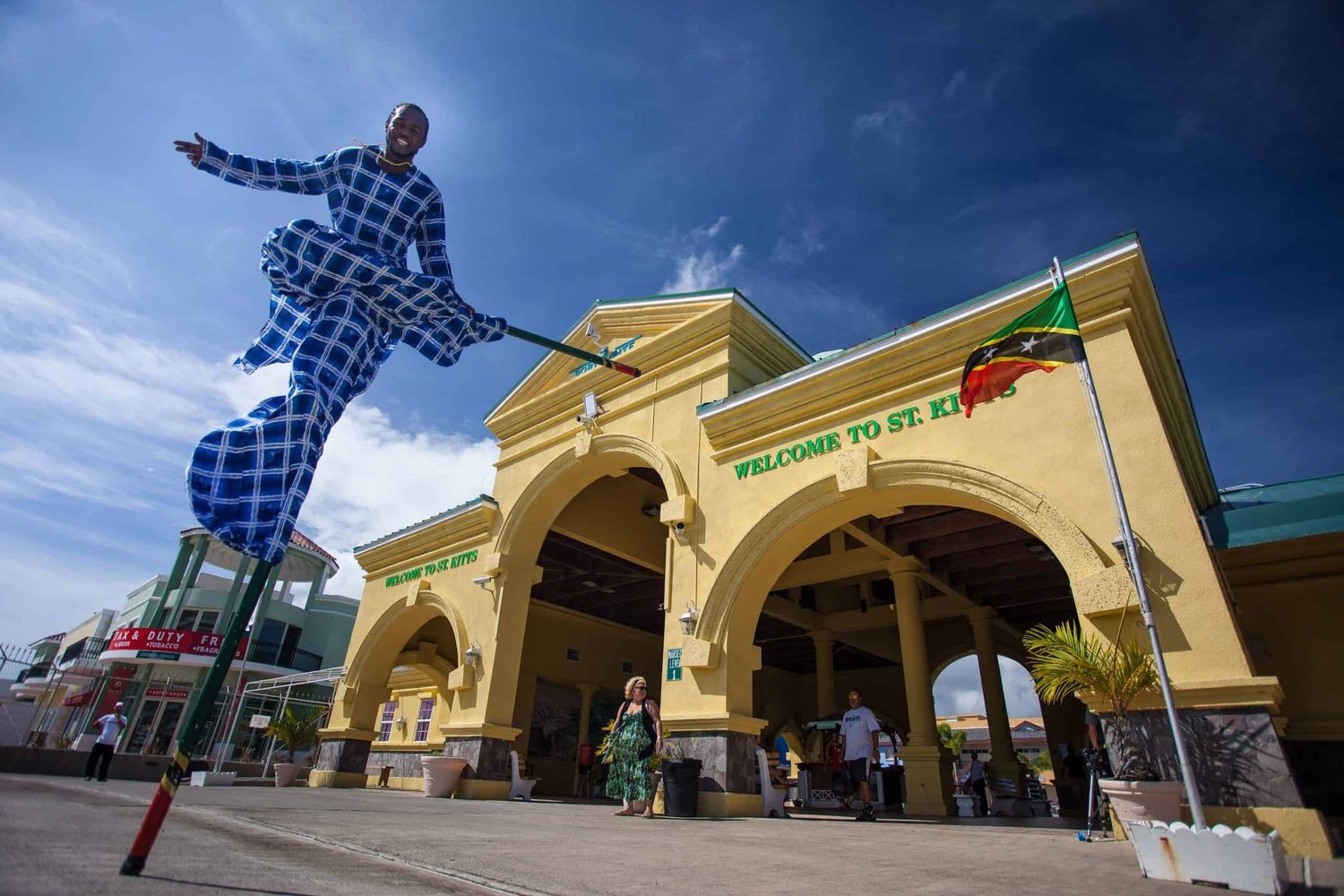 Stilt walker performer in blue outfit at Welcome to St. Kitts port entrance under dramatic cloudy sky.