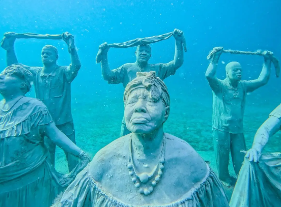 Underwater sculptures of human figures holding fabric above water, covered in algae, in a blue ocean setting.