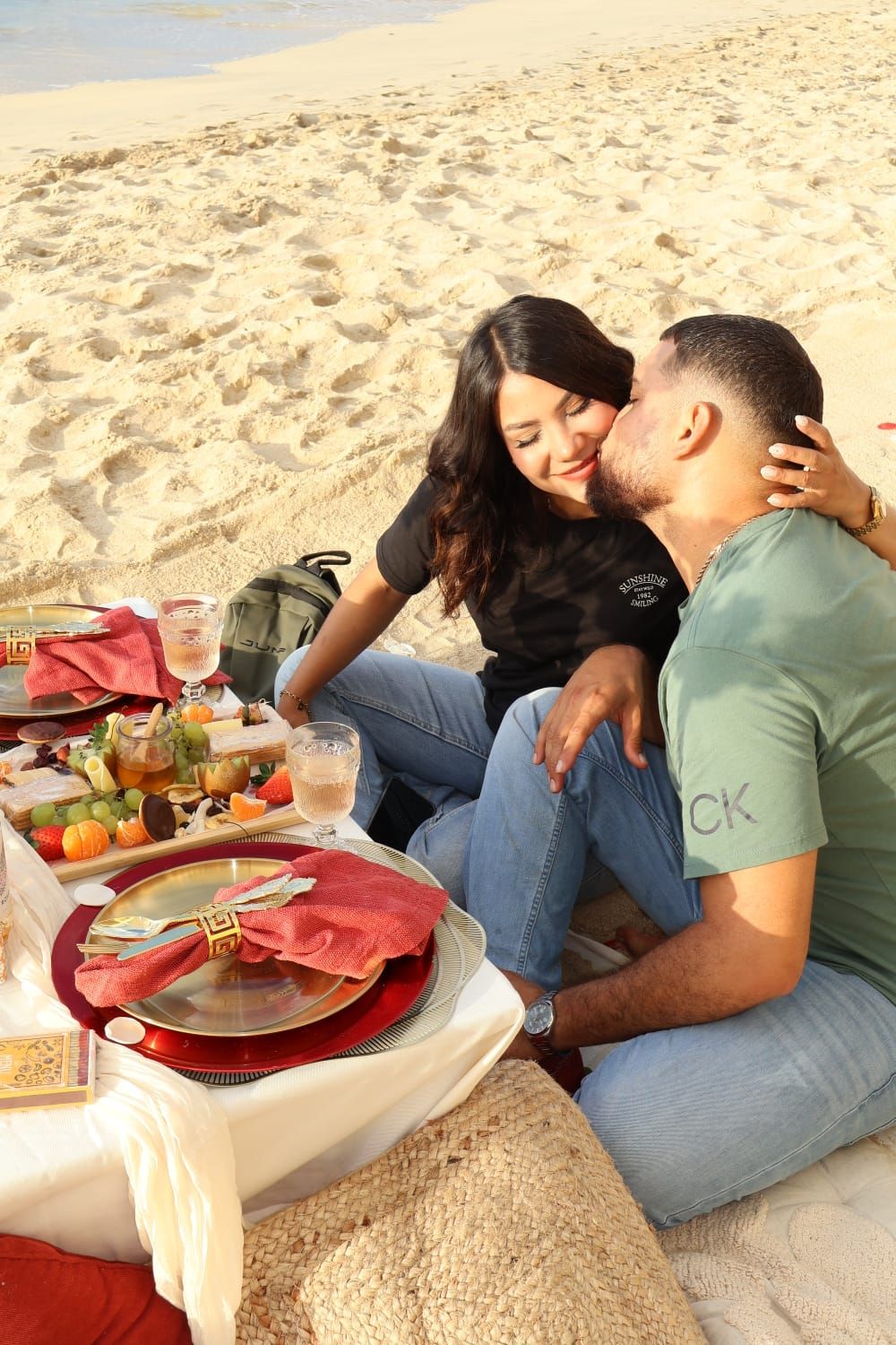 Couple sharing a romantic beach picnic with gourmet food and elegant place settings on a sunny day.