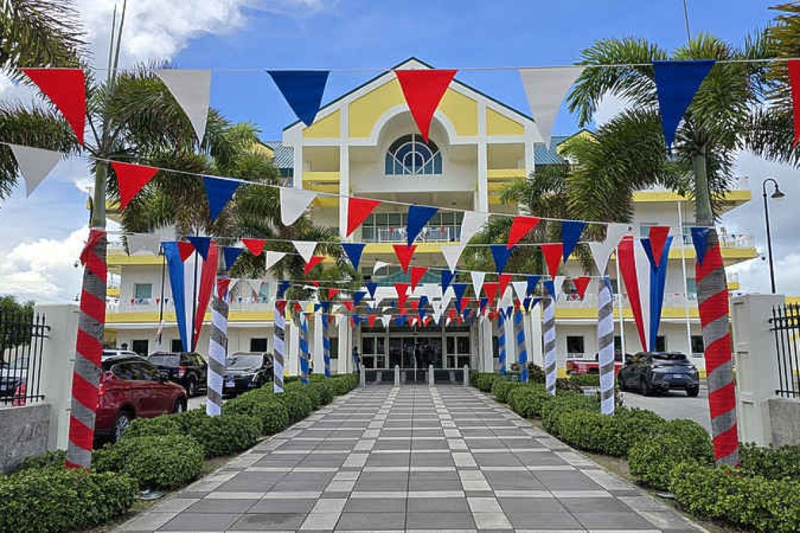 Colorful bunting decorates a yellow government building entrance flanked by palm trees and red-striped bollards.