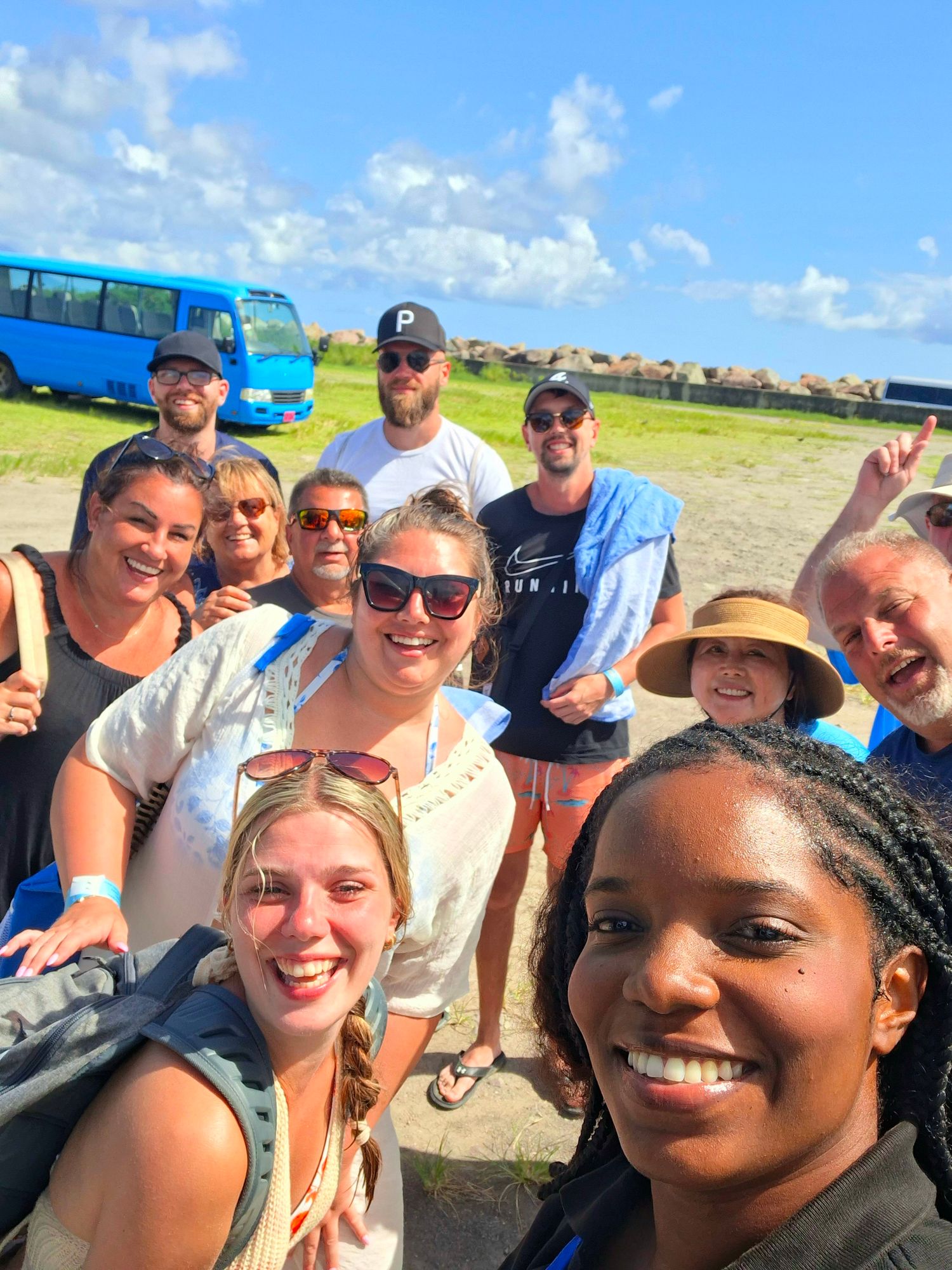 Group of smiling tourists taking a selfie outdoors near a blue bus on a sunny day during a sightseeing tour.