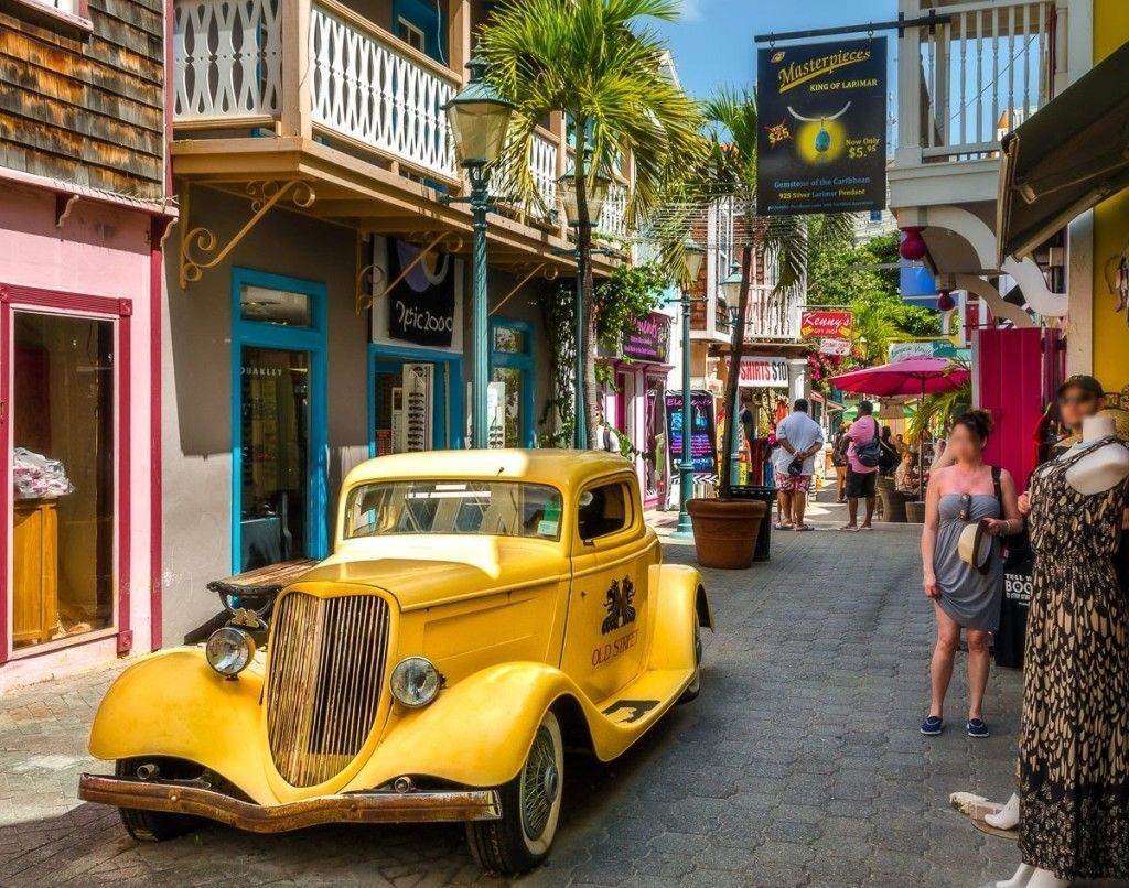 Vintage yellow classic car parked on a colorful pedestrian shopping street with tourists and tropical palm trees.