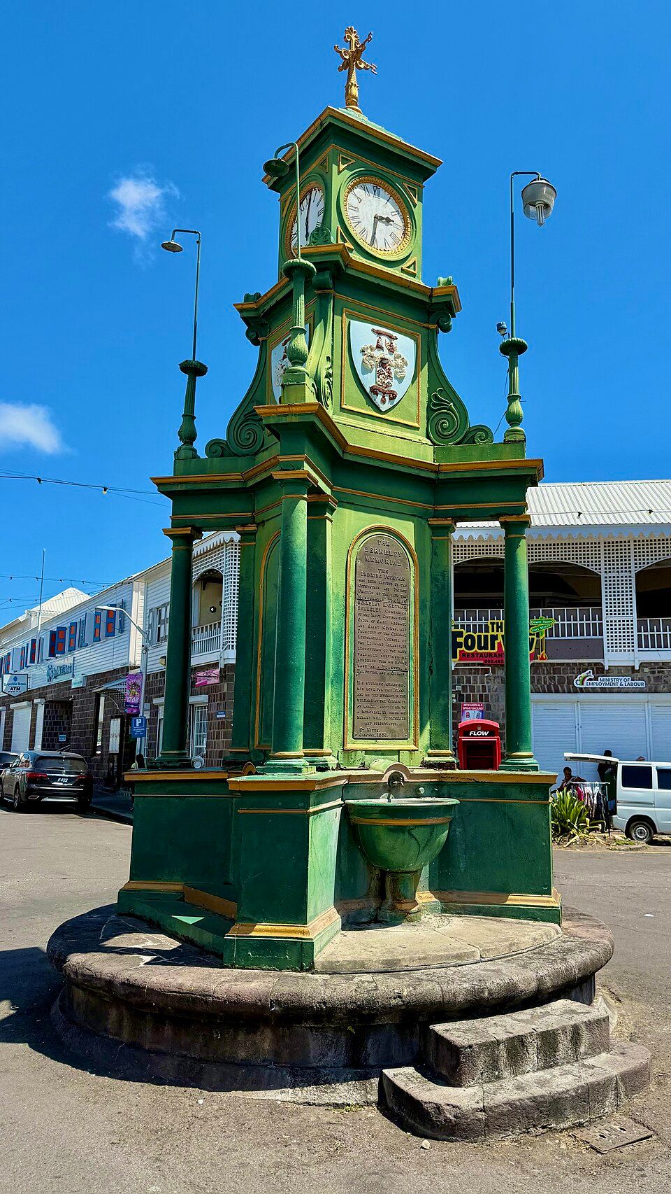 Green Victorian clock tower with coat of arms stands in Basseterre, Saint Kitts town square under bright blue sky.